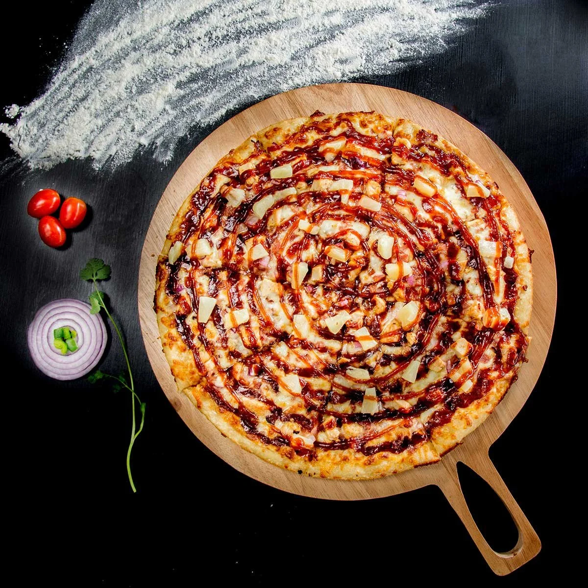 A pepperoni pizza with cheese on a round wooden pizza peel, garnished with diced onions, with cherry tomatoes, sliced onion, and green herbs on a black surface.