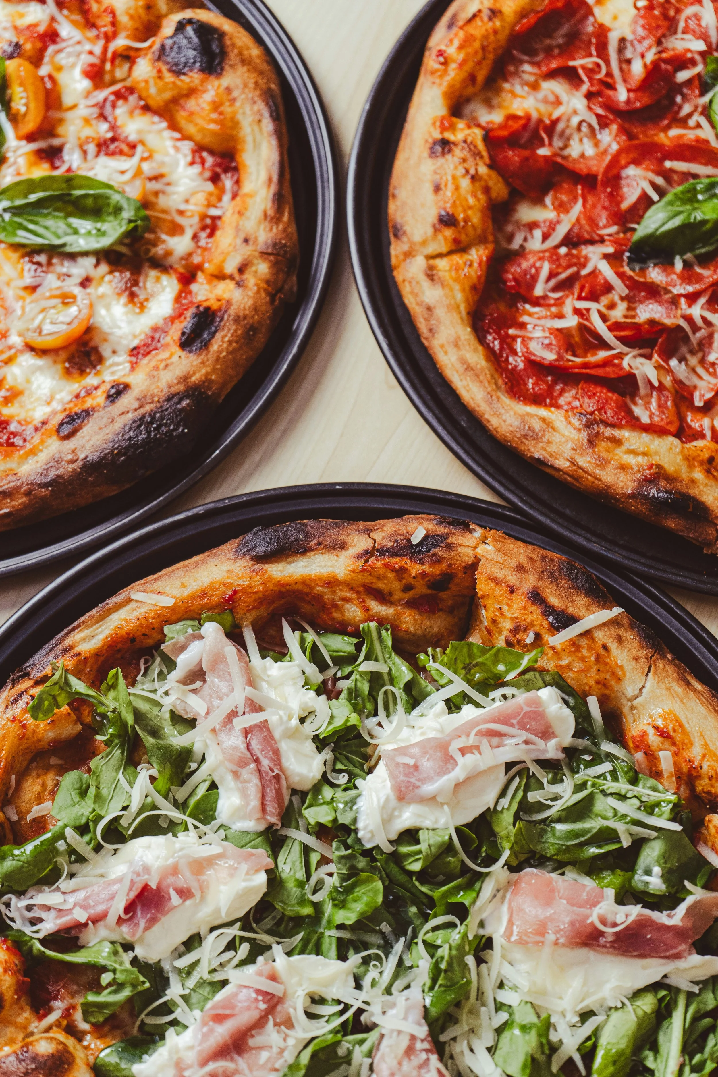 Three pizzas in black pans on a wooden table, one with basil and cheese, another with tomato and cheese, and the third with arugula, prosciutto, and cheese.
