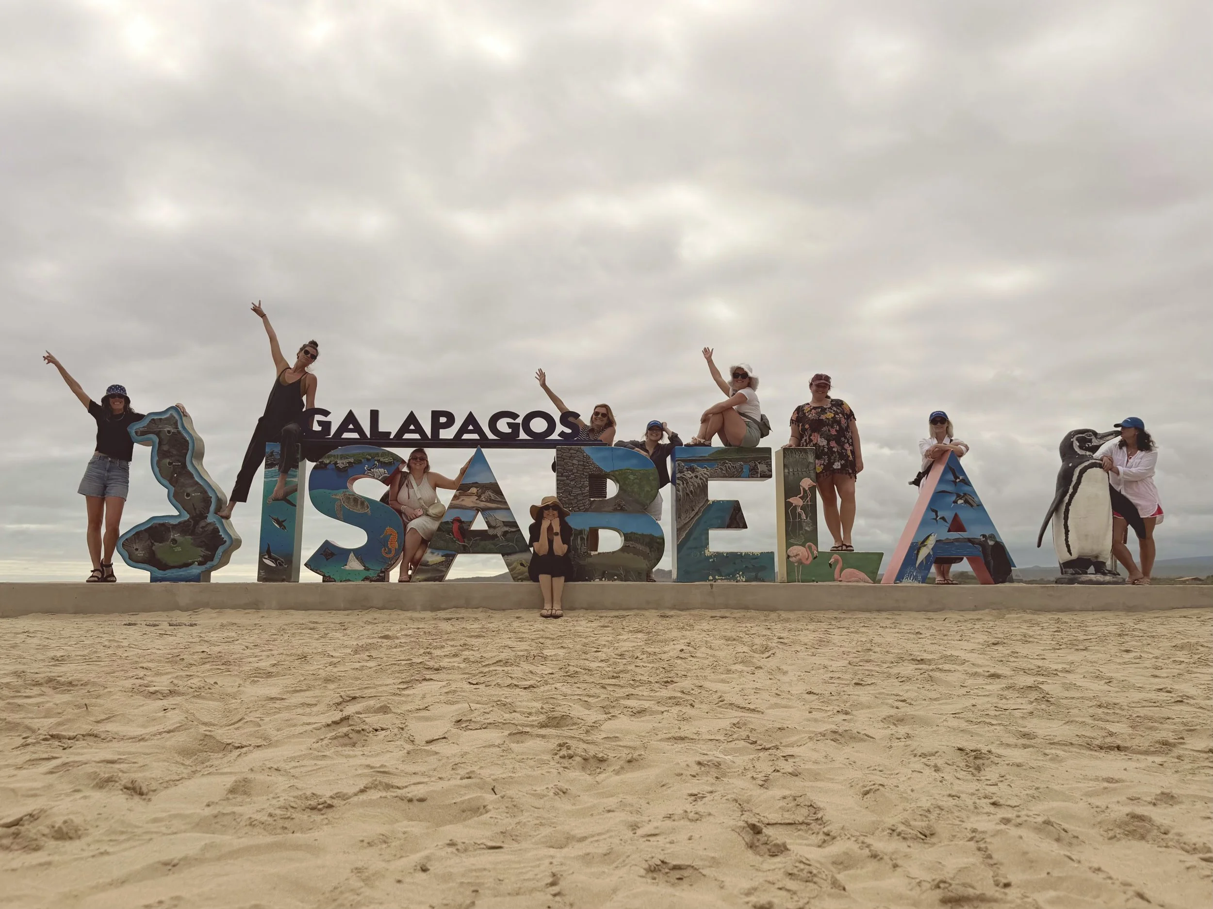 Group of people posing with large colorful Galápagos Islands sign on a sandy beach.
