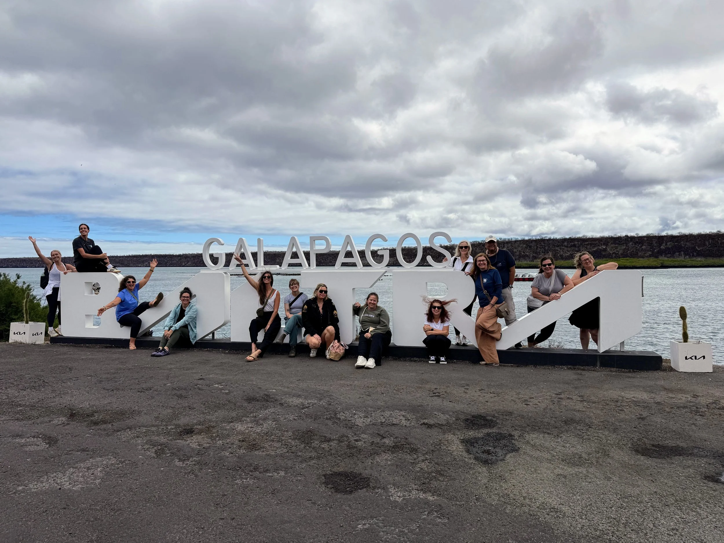Group of people posing with large white sign that reads 'Galapagos' and 'Ecuador' against a cloudy sky and water background.