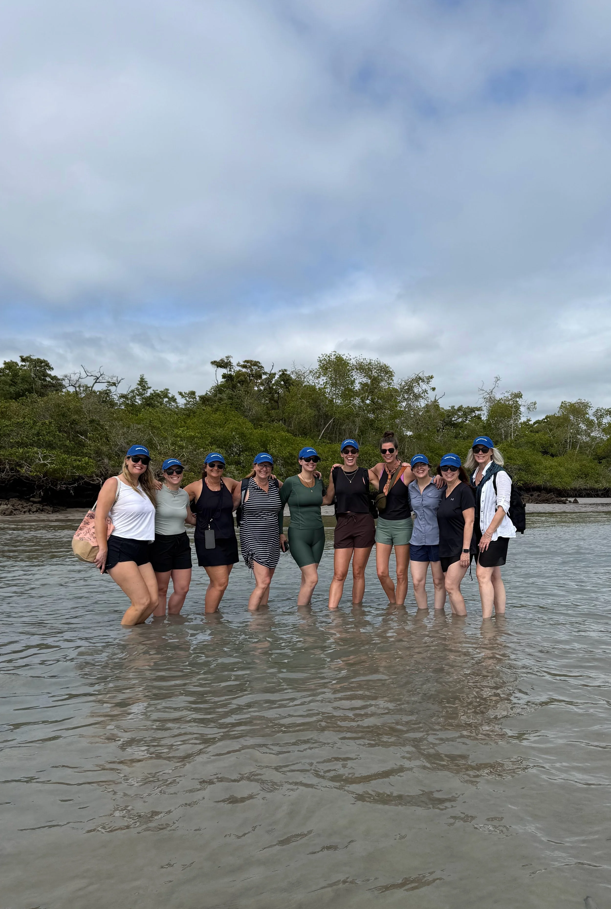 Group of women standing in shallow water at the beach, wearing casual summer clothes and blue caps, with trees and cloudy sky in the background.