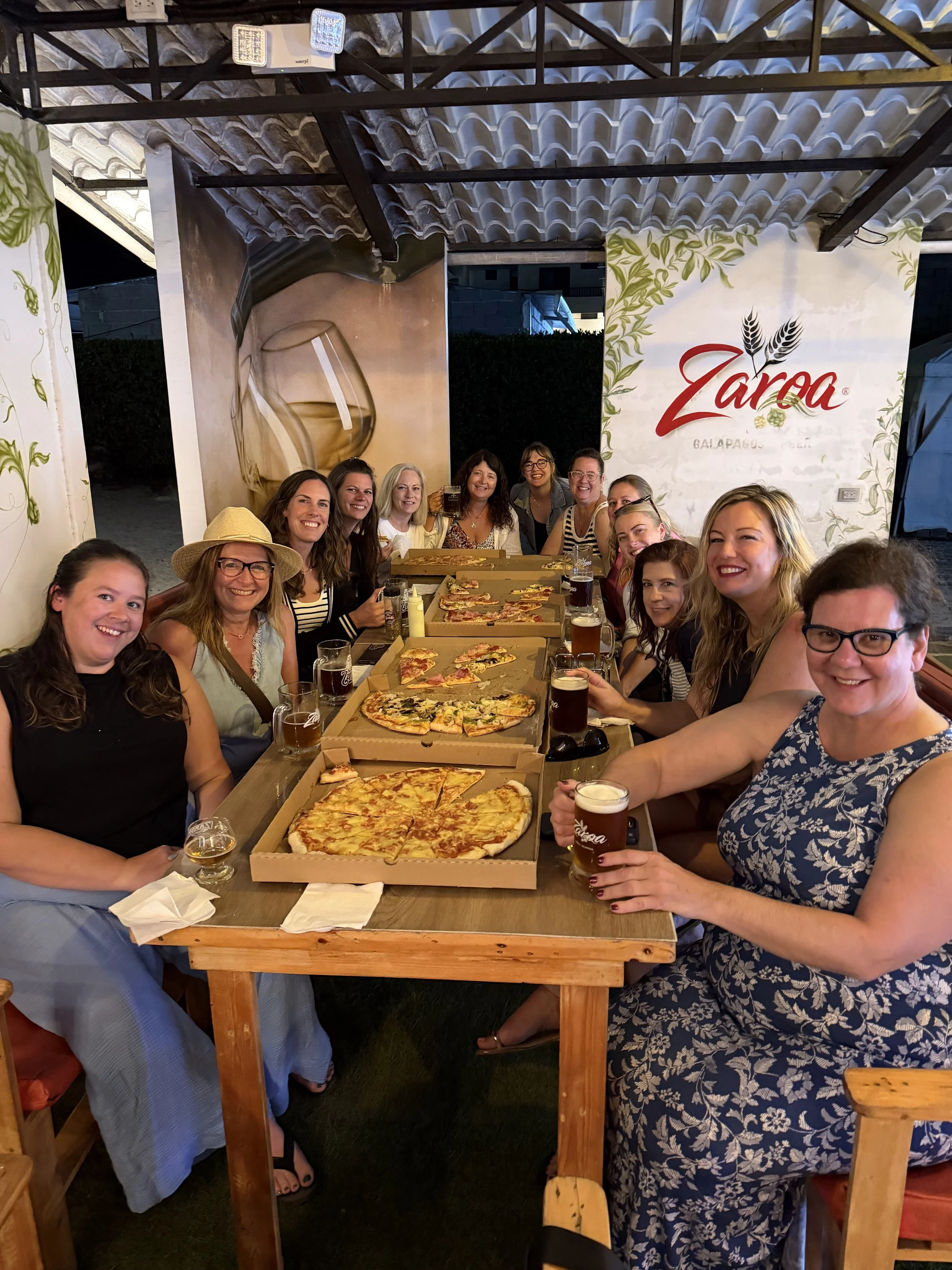 Group of women gathered around a wooden table with pizza boxes and drinks in a restaurant or bar setting, smiling at the camera.