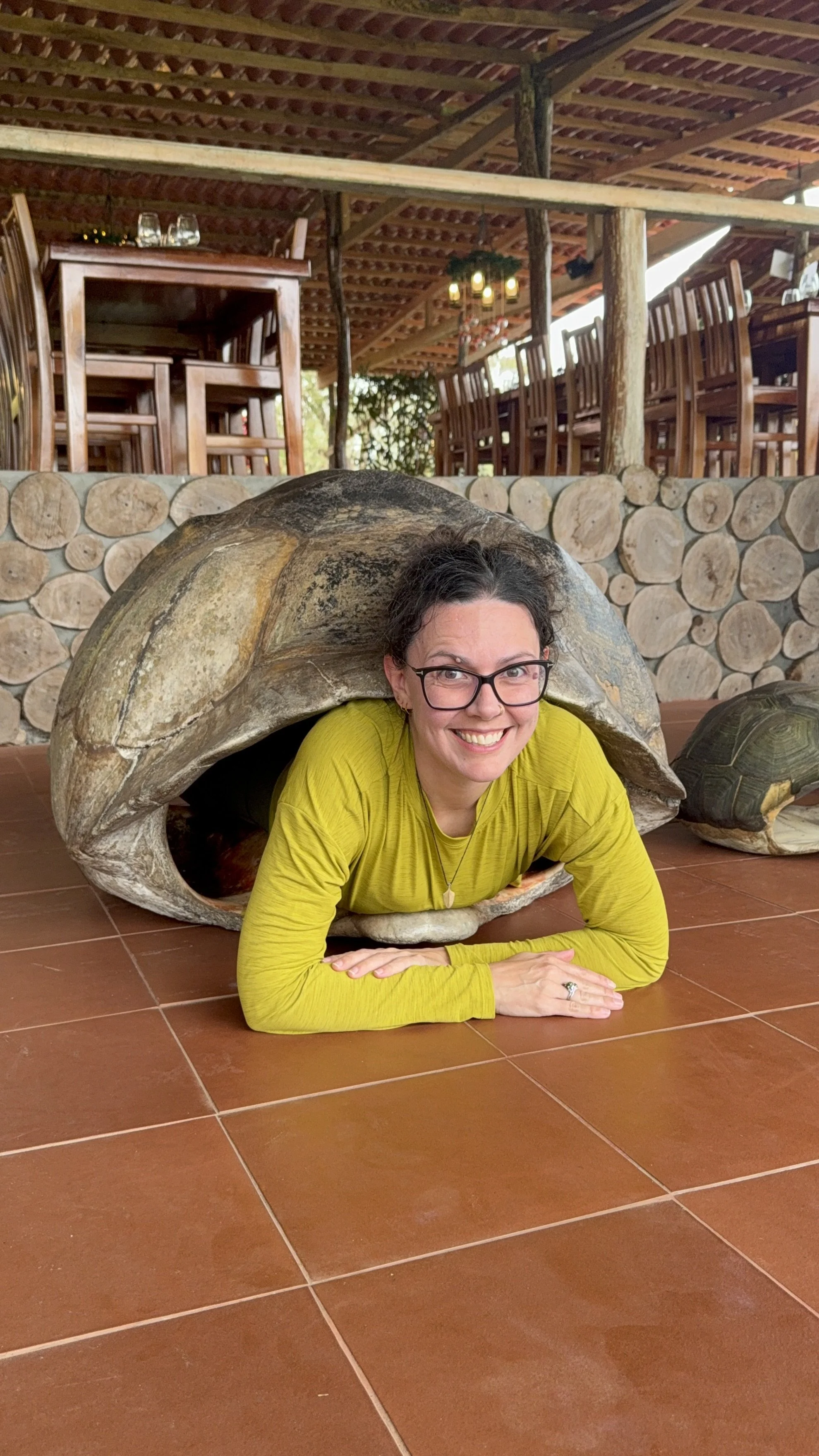 Woman smiling and laying on the floor inside a turtle shell, with wooden furniture and logs in the background.