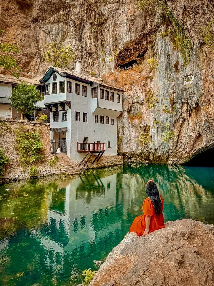 A widowed woman sitting on a rock by a river, with a house built into a rocky cliff in the background, taking time for herself and the beauty around her. 