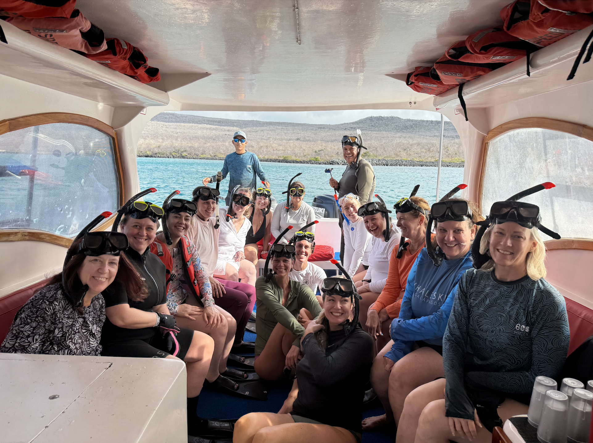 Group of people on a boat preparing for scuba diving, wearing wetsuits and snorkeling masks with snorkels, with a lake and mountains in the background.
