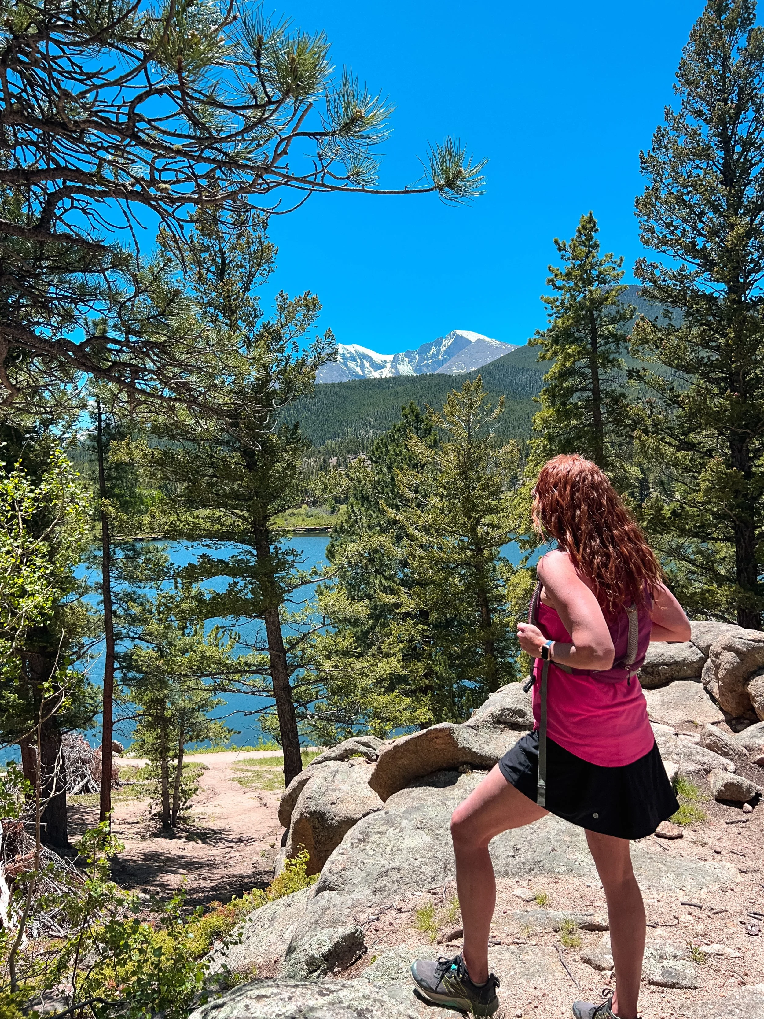 A young adventurous widowed woman with curly red hair wearing a pink tank top, black shorts, and hiking shoes stands on a rocky trail, looking at a mountain landscape featuring pine trees, a lake, and snow-capped peaks under a bright blue sky.