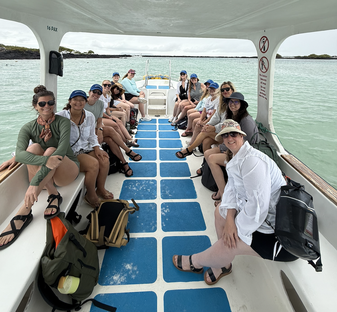A group of women sitting on benches and floor of a boat, wearing casual clothes and hats, with backpacks nearby, with water and distant land in the background.