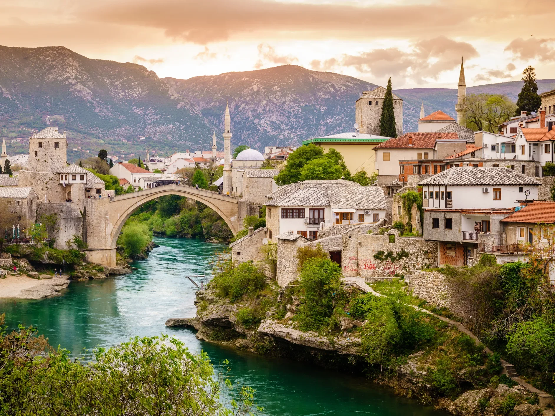 A scenic view of a historic city with a stone bridge over a turquoise river, surrounded by old buildings, greenery, and mountains in the background during sunset.