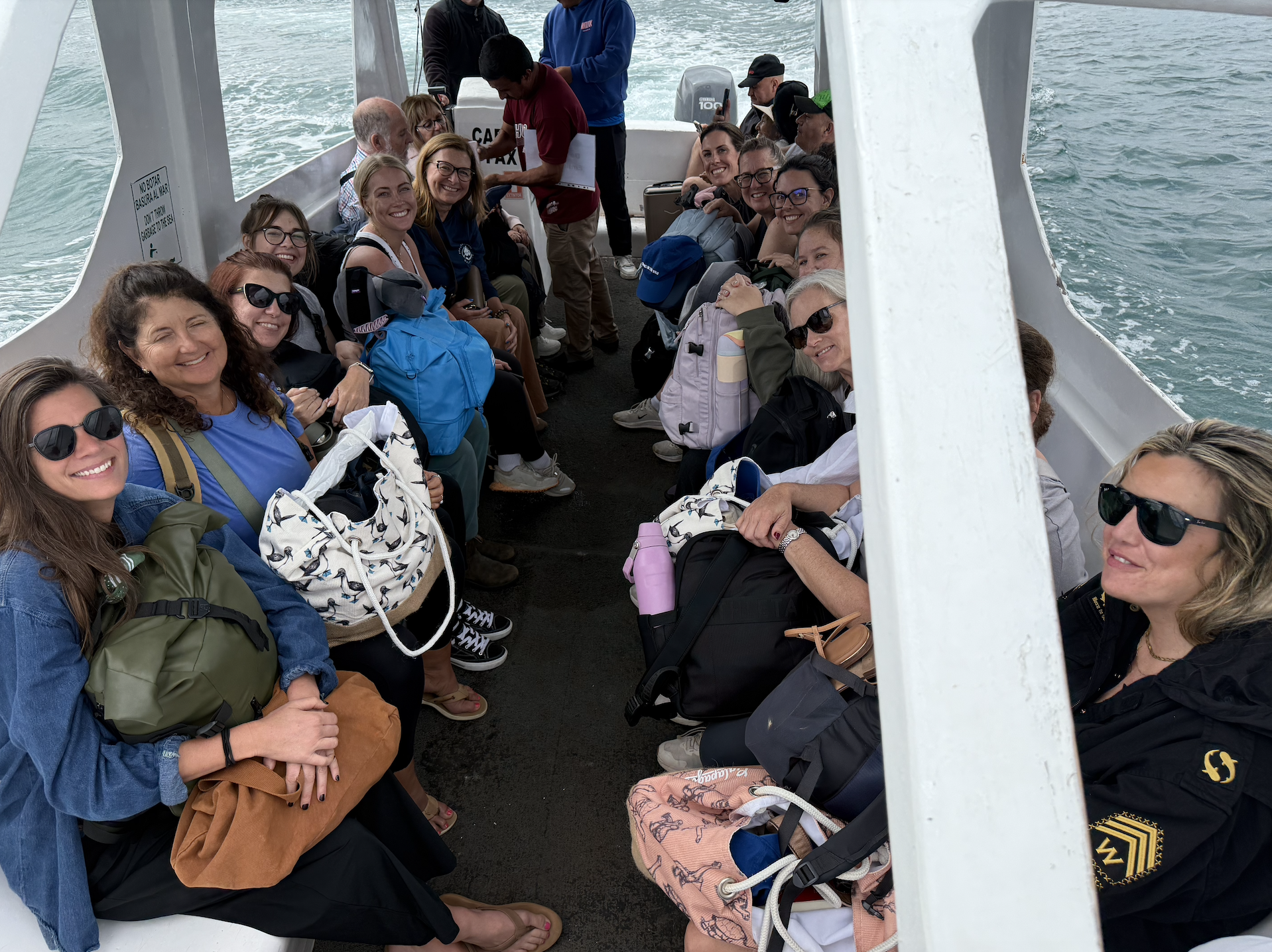 A group of smiling women sitting on a ferry boat, carrying backpacks and sunglasses, with water visible on both sides of the boat.