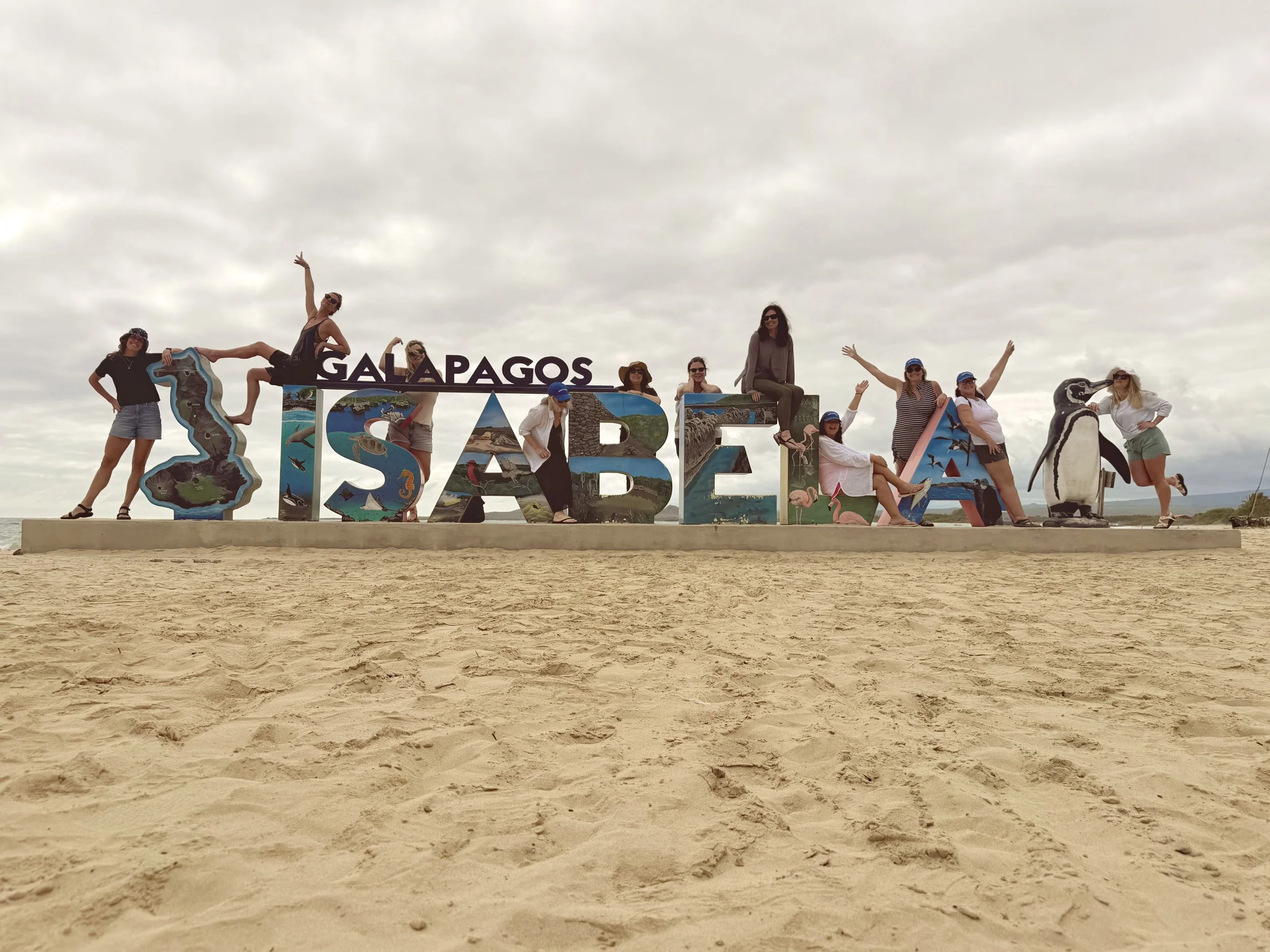 Group of people standing and posing around colorful sign that reads 'Galápagos ISABELA', with a penguin figure on the far right, on sandy ground under cloudy sky.