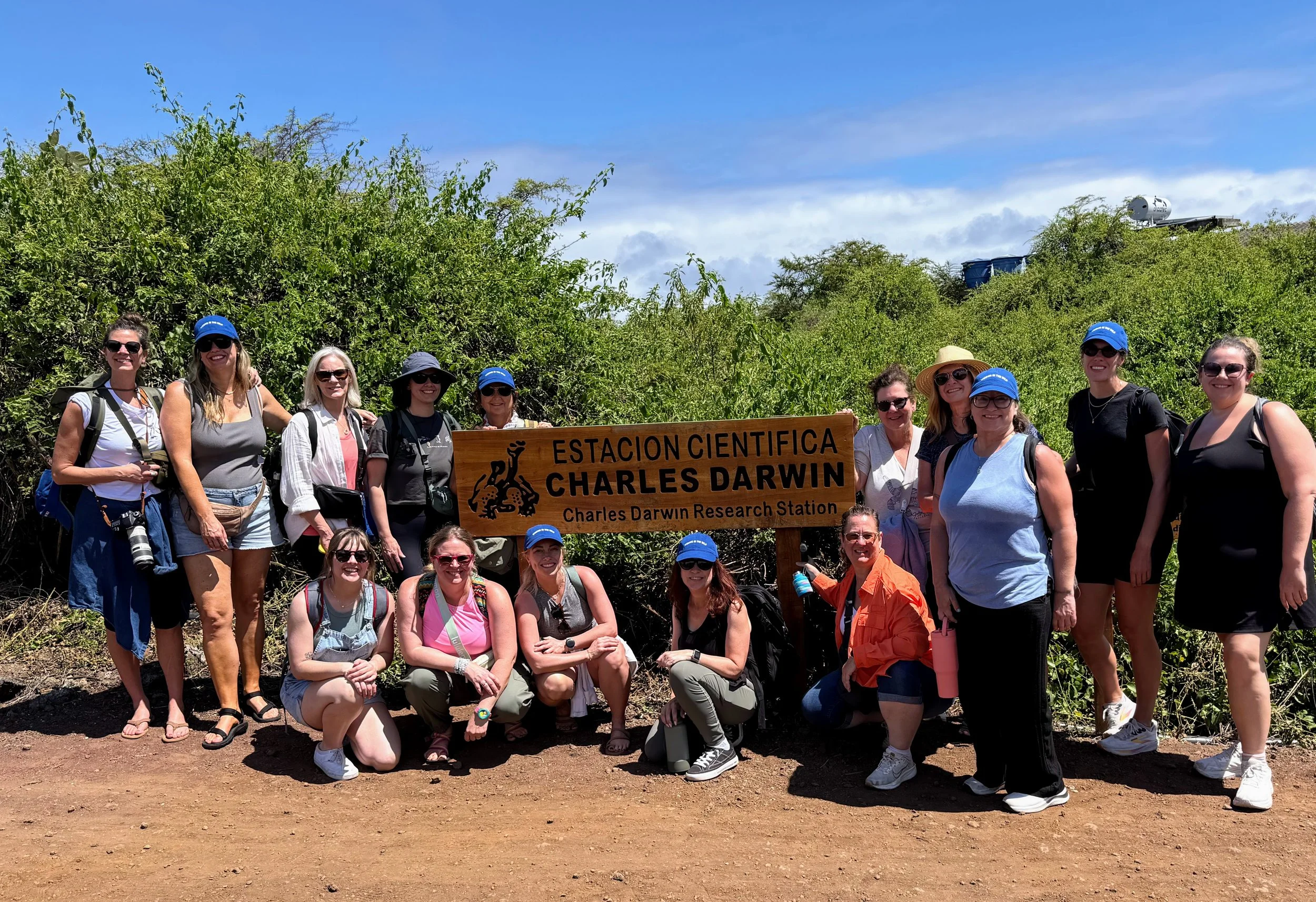 Group of people on a hiking trail holding a wooden sign that reads 'Estacion Cientifica Charles Darwin, Charles Darwin Research Station' in front of green bushes and blue sky.