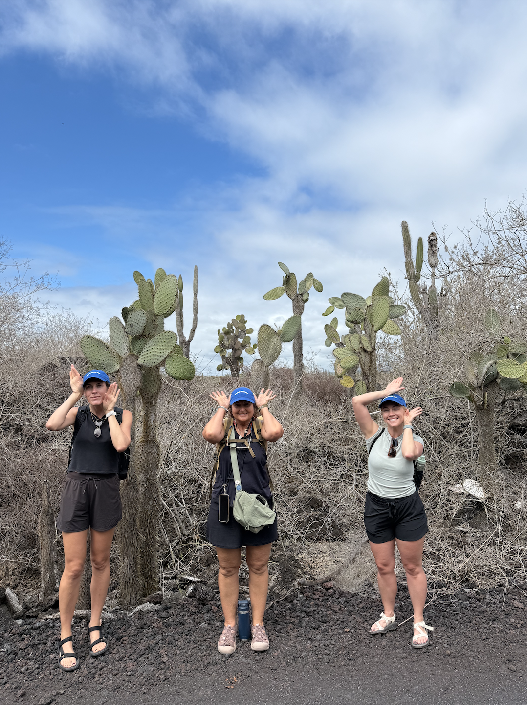 Three women with backpacks and blue caps standing in front of desert cacti and dry bushes, under a partly cloudy sky.