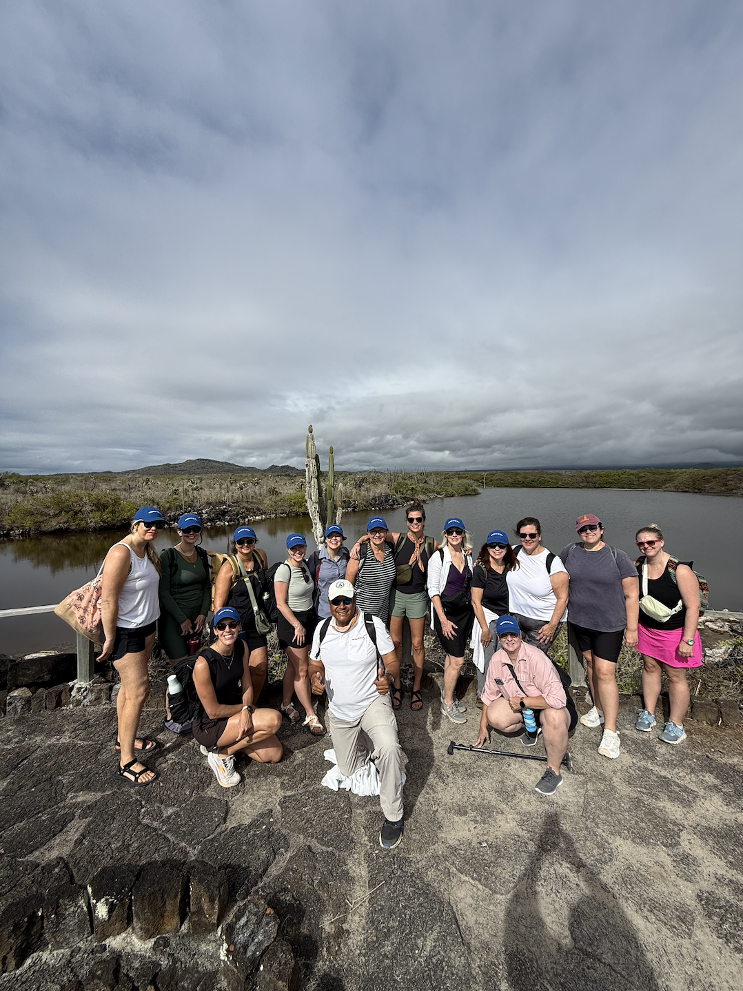Group of people hiking outdoors next to a lake with desert vegetation and cacti, cloudy sky.