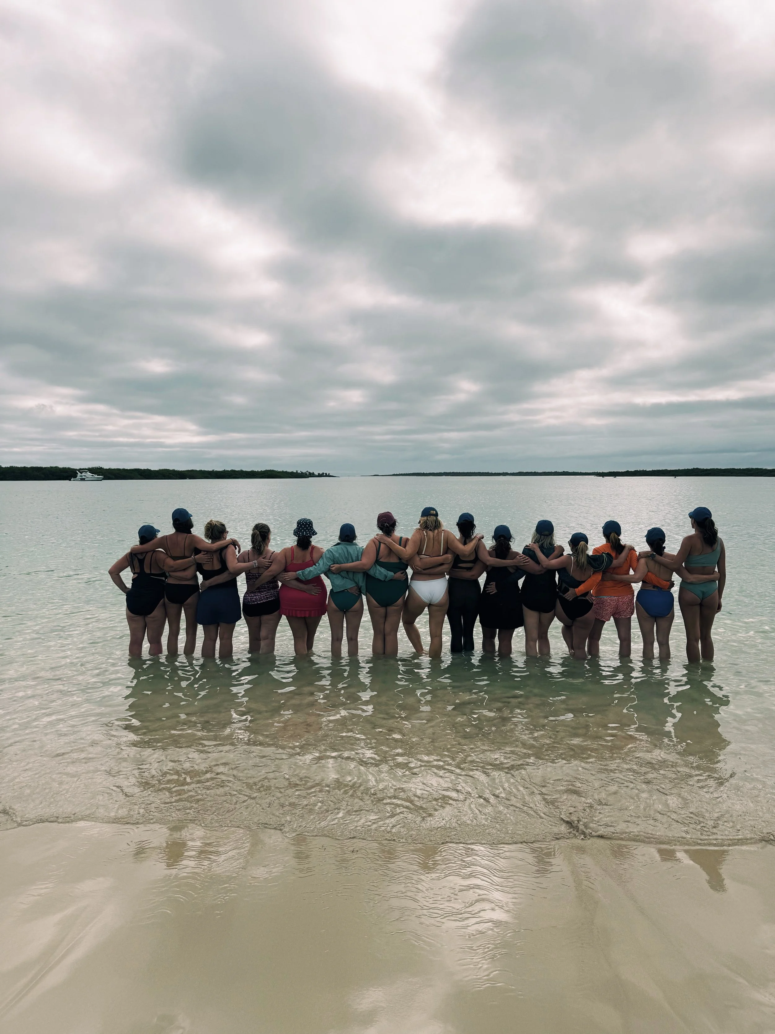 A group of women standing in the water on a beach, arm in arm, facing the ocean under a cloudy sky.