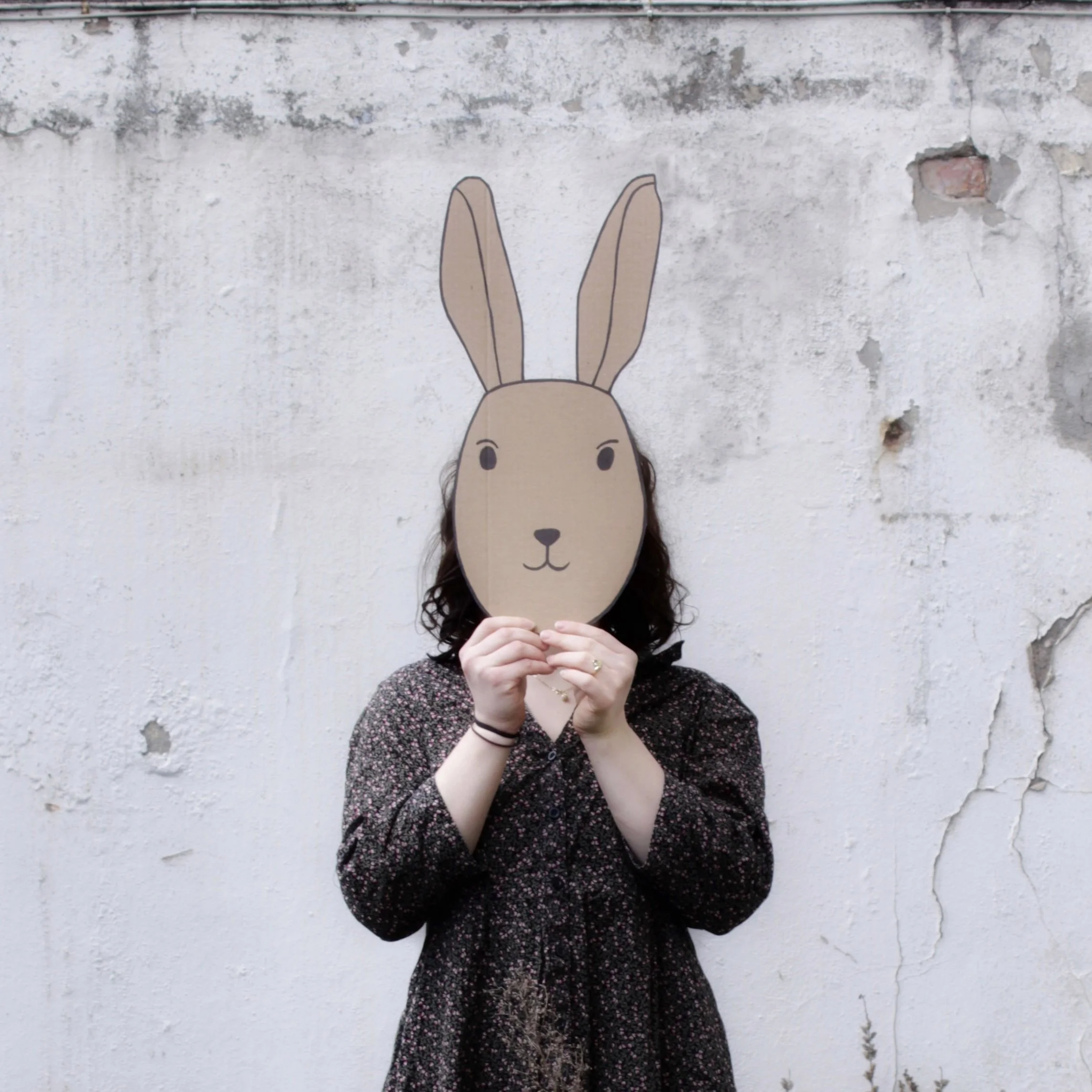 Frau mit dunklen Locken hält sich eine Hasen Maske aus Karton vor das Gesicht.