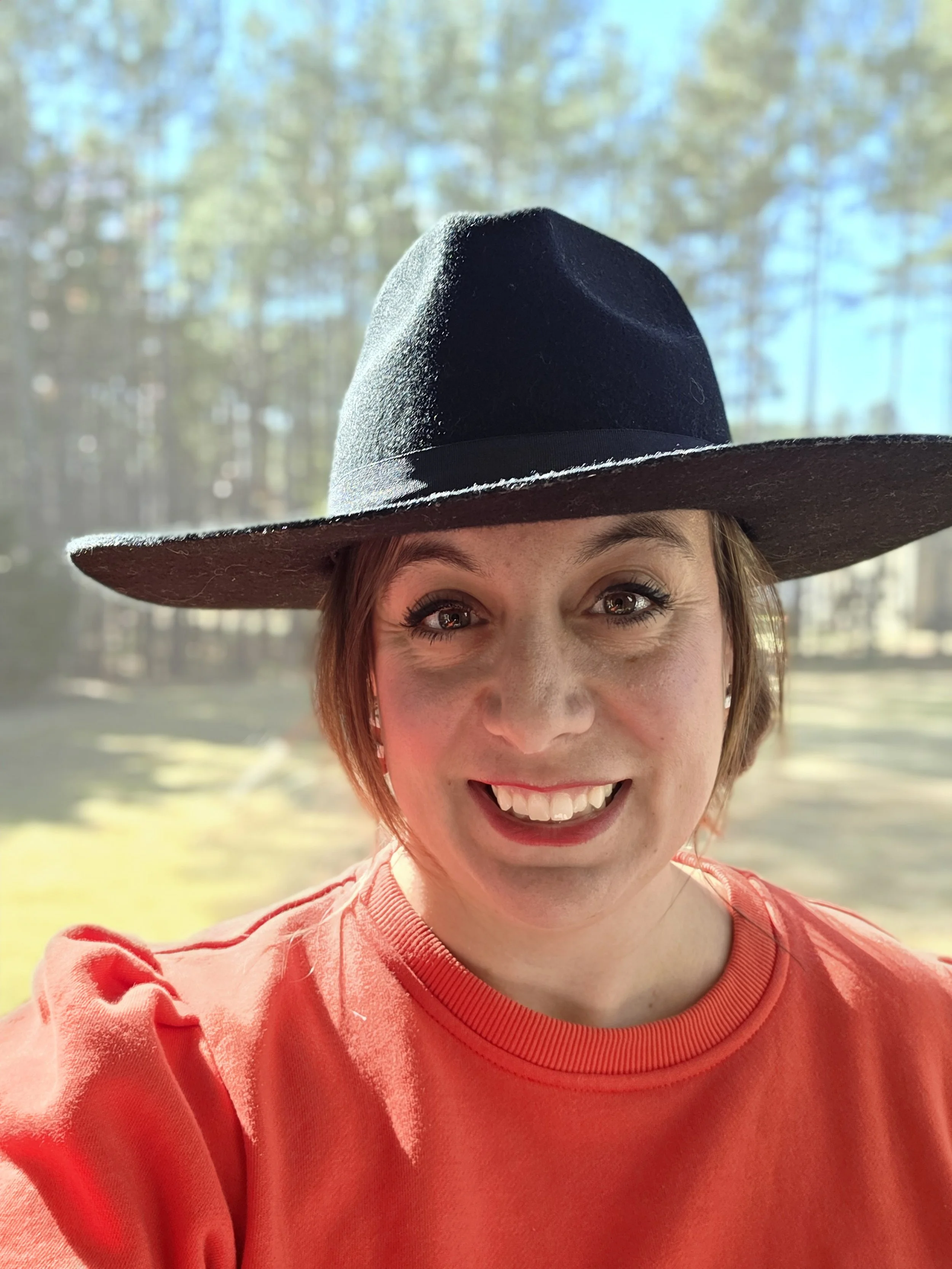 A woman with short hair wearing a wide-brimmed black hat and a red shirt, smiling outdoors in front of a forest with sunlight filtering through trees.