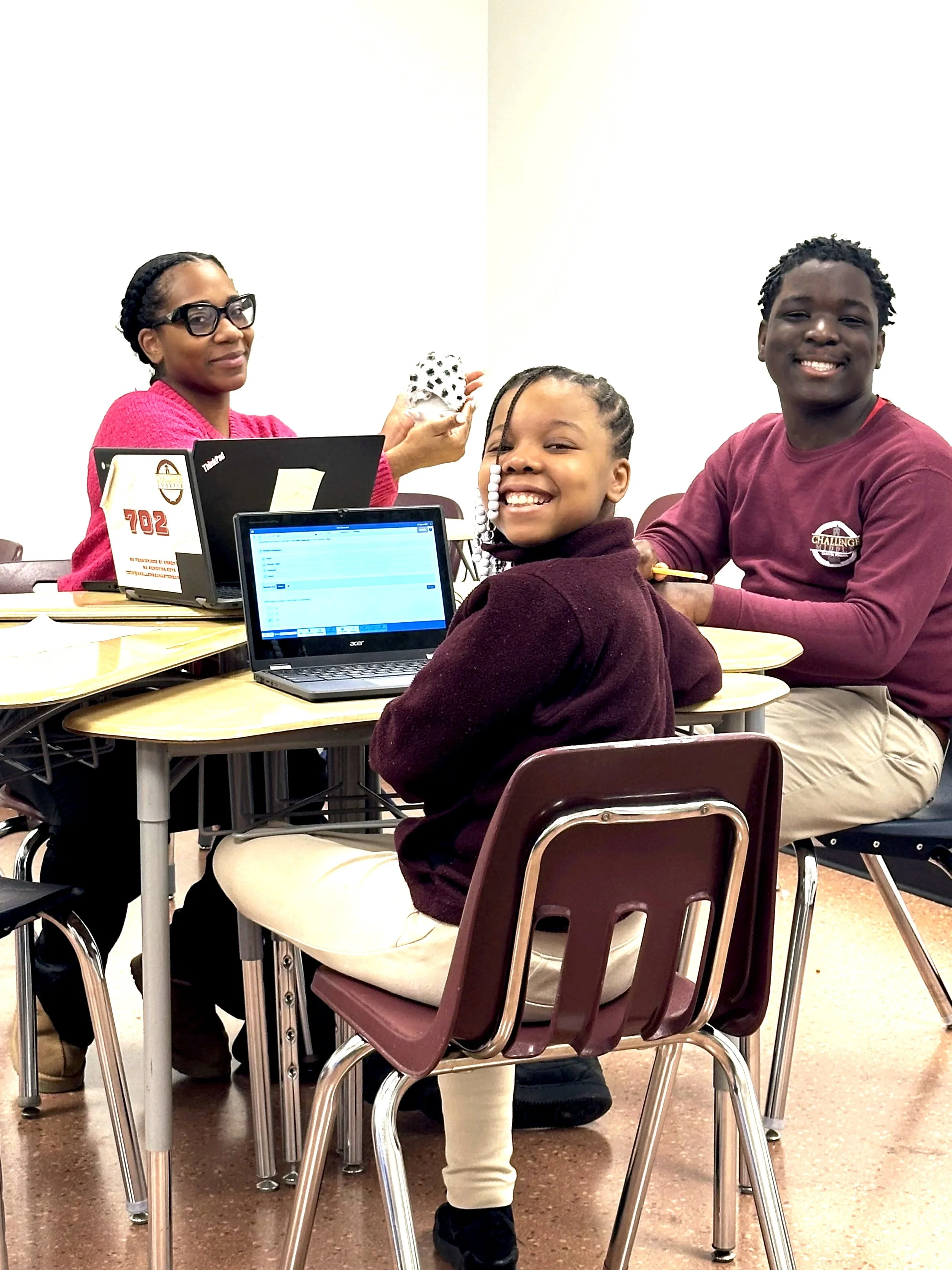 A teacher and two scholars wearing maroon shirts and big smiles are sitting at a classroom table