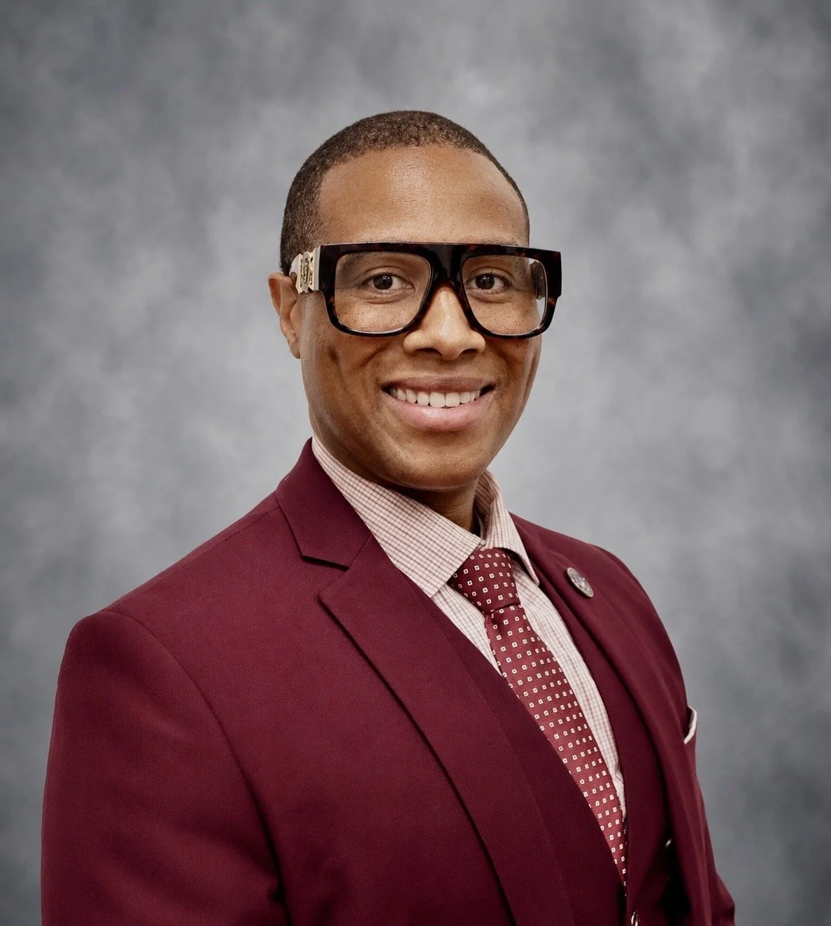 picture of a man with glasses wearing a maroon suit jacket and tie
