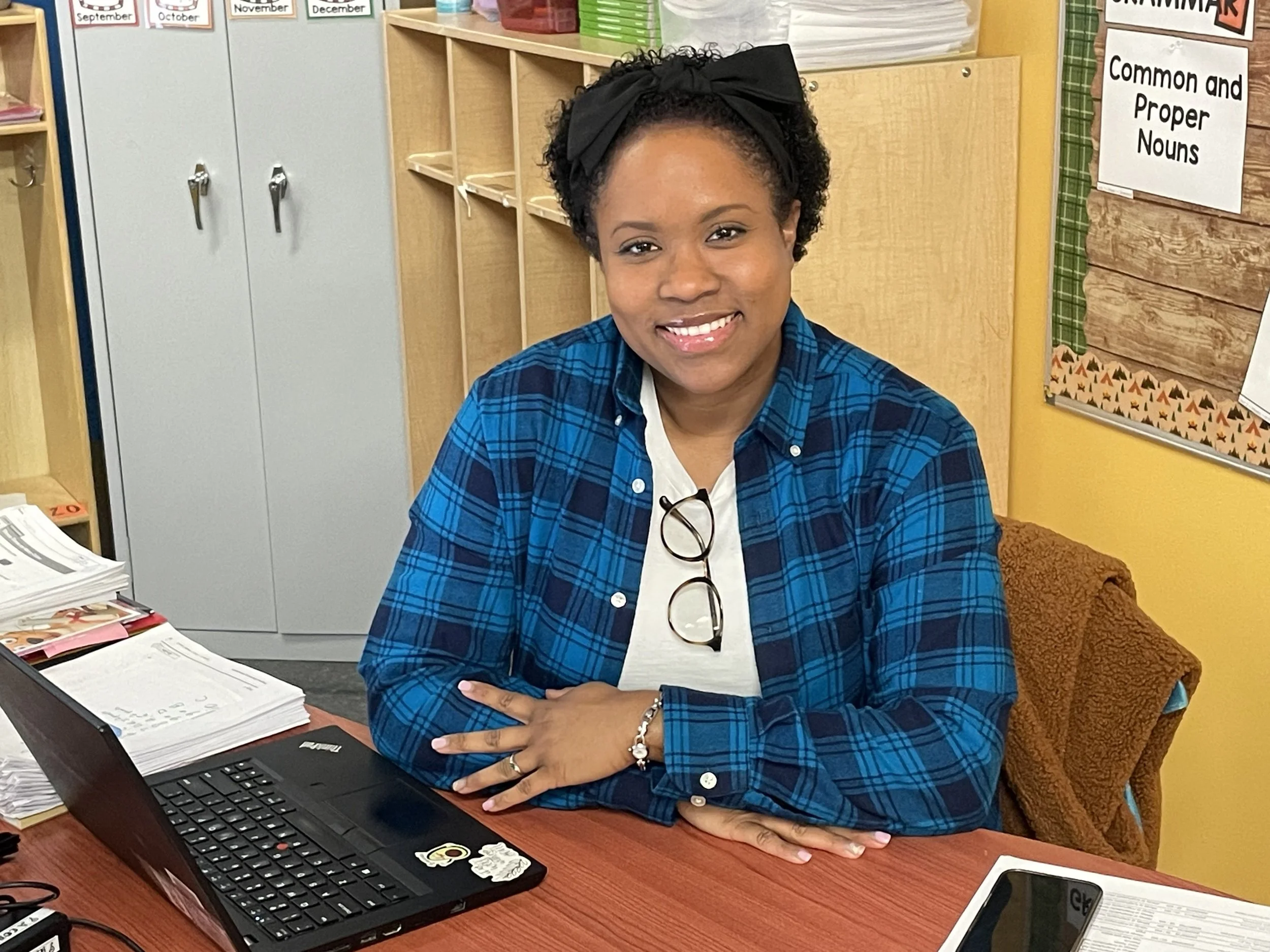 a teacher in a blue and black flannel shirt sits in her classroom smiling at the camera