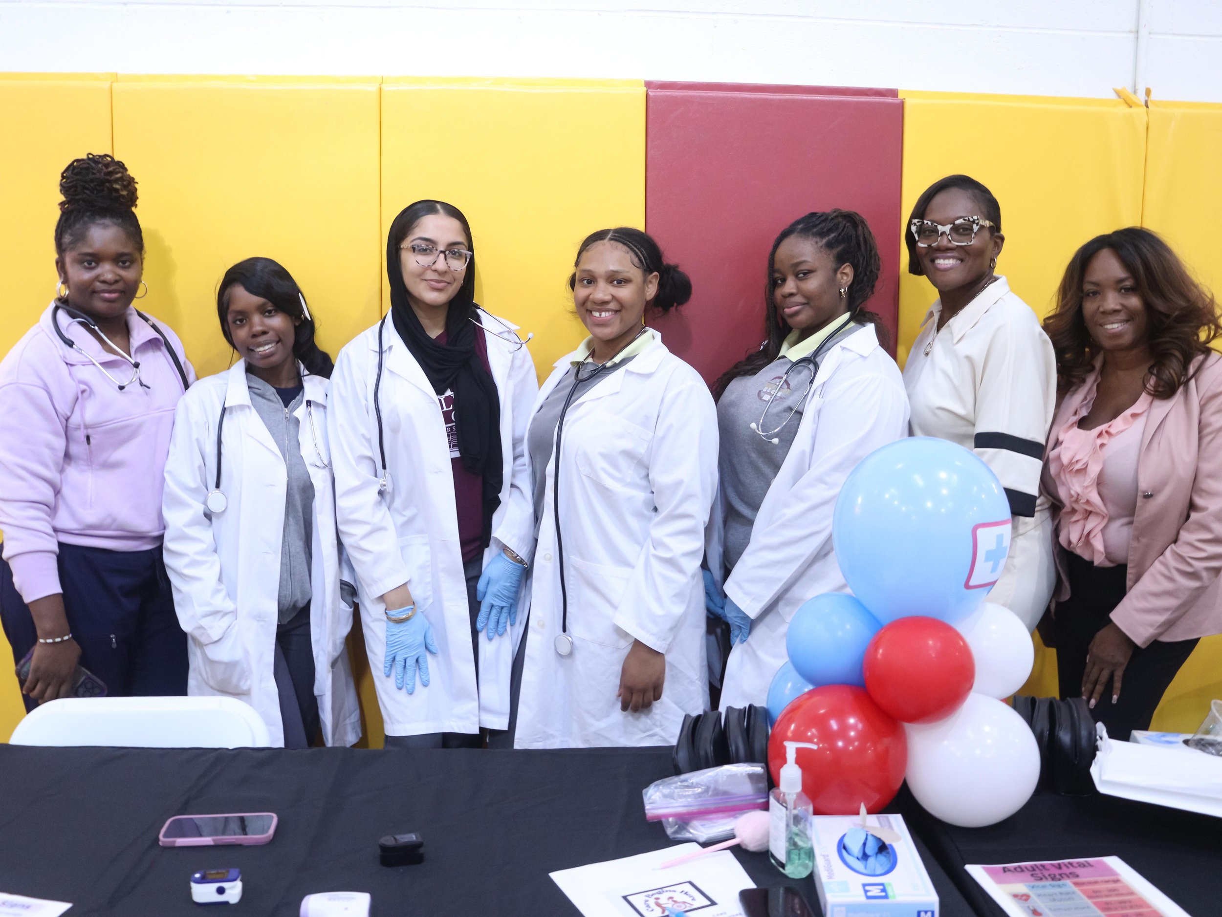 several female scholars stand with medical coats and stethoscopes around their neck at a community health fair
