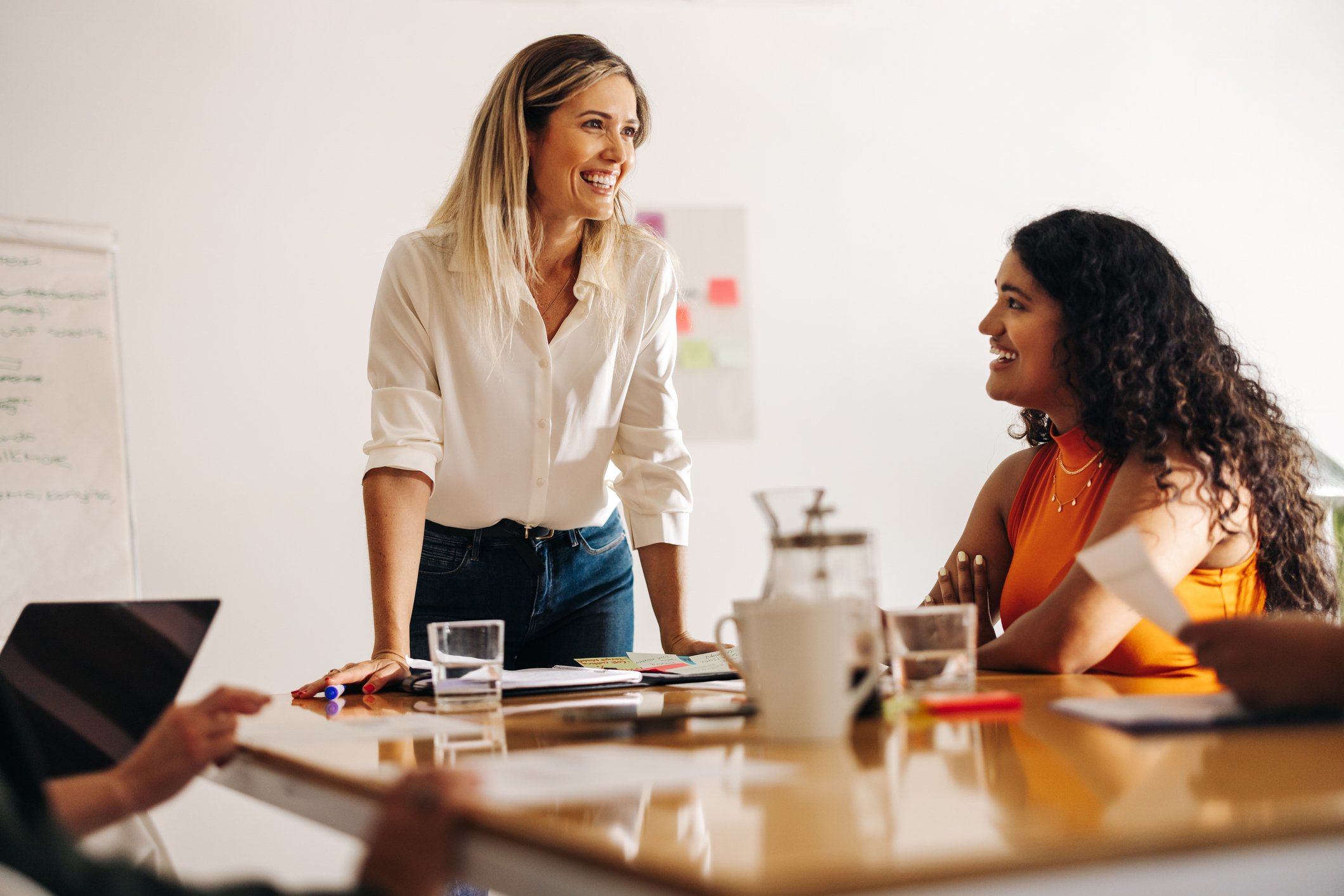 Two women smiling and talking in a meeting room with papers, notebooks, and drinks on the table.