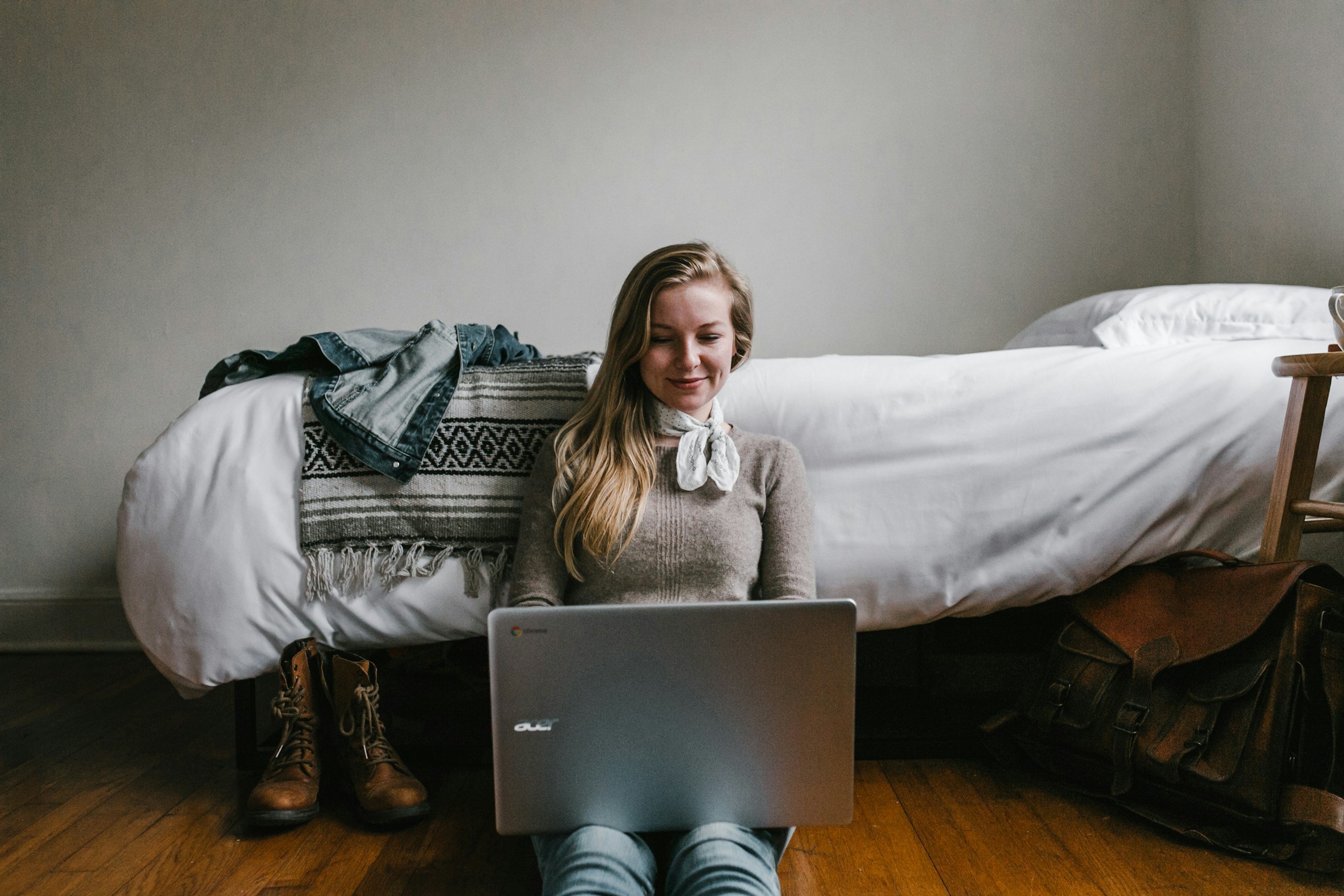A young woman sitting on the floor in front of a bed, using a laptop, with boots and a bag nearby.