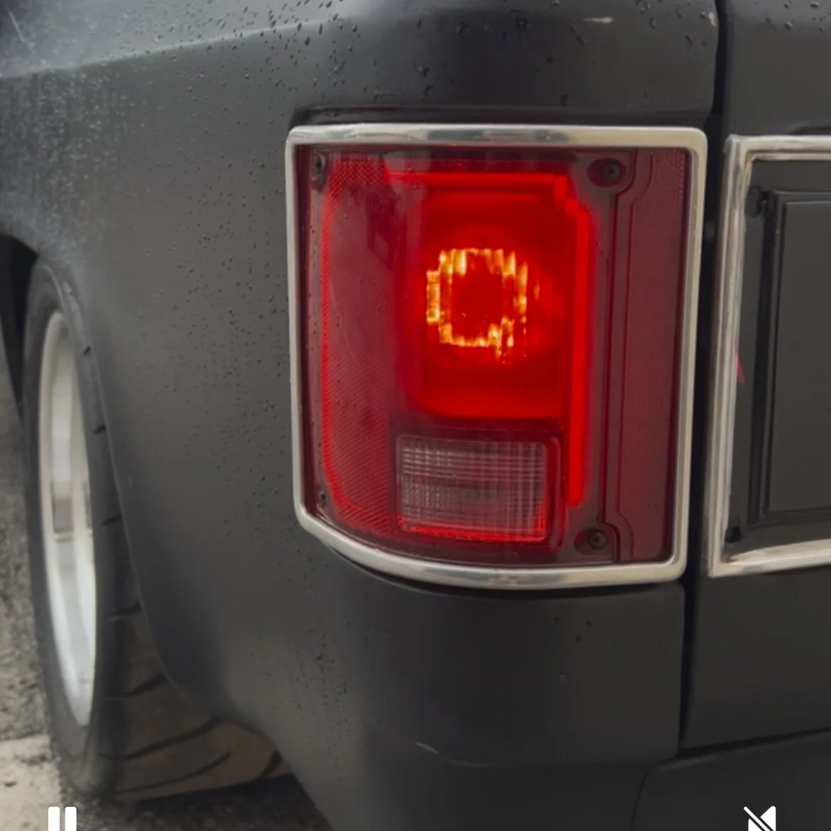 Close-up of the rear tail light of a black vehicle, with the brake light illuminated and rain droplets on the car's surface.