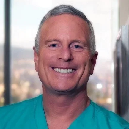 Portrait of a smiling man with short gray hair, wearing a teal medical scrub, standing indoors near a window.