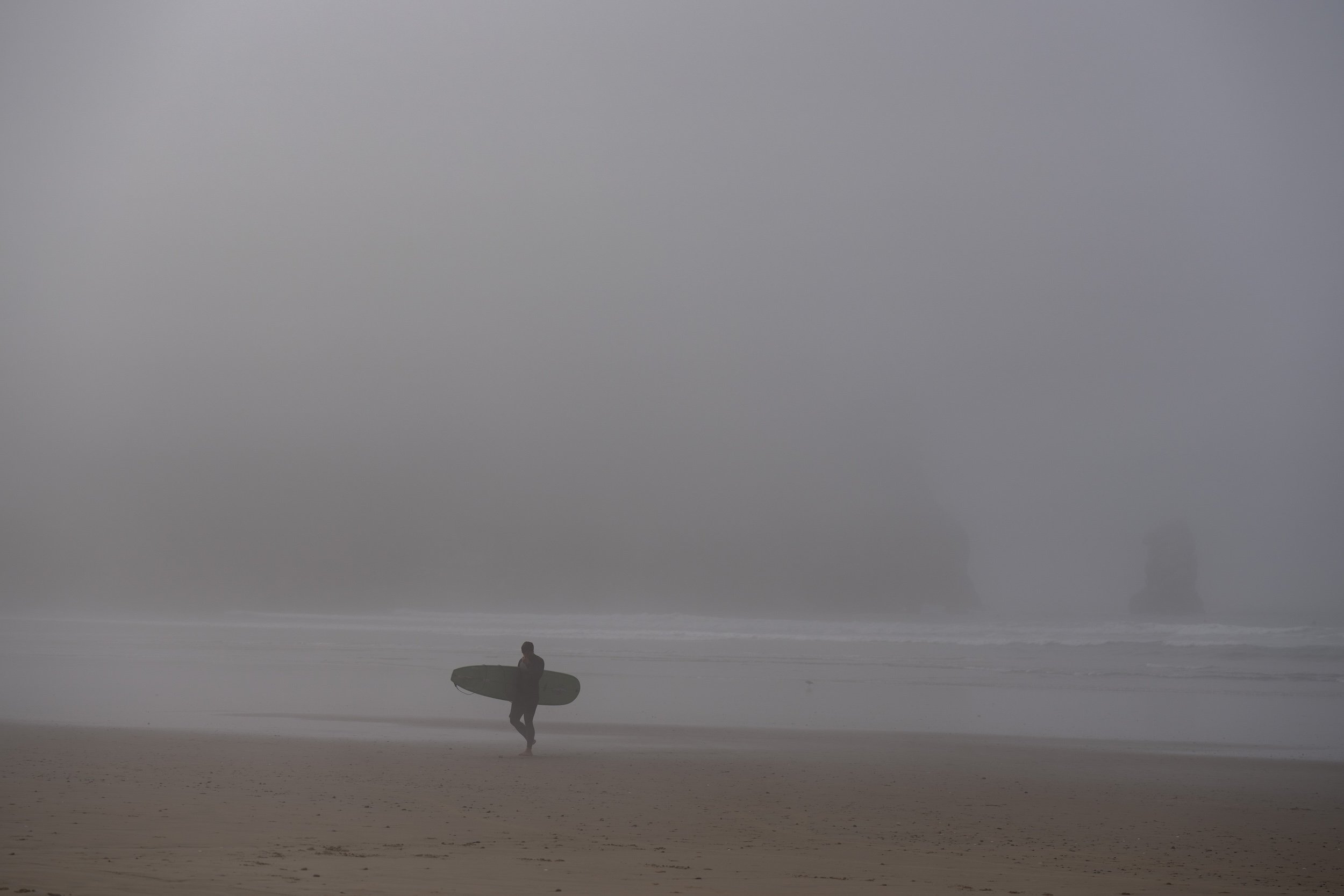Person walking a dog across a foggy beach with reflections on wet sand and the ocean in the distance.