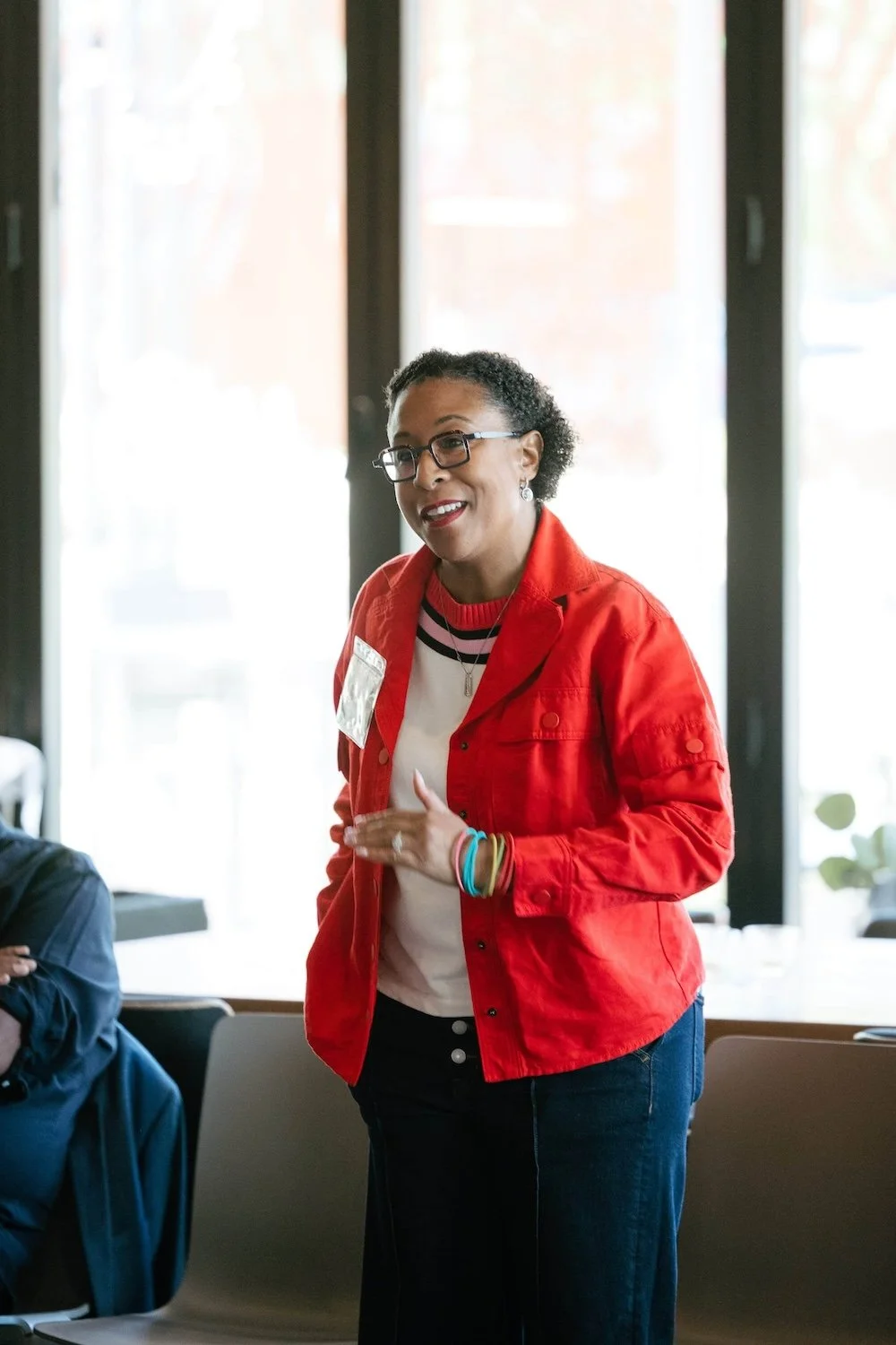 A woman in a red jacket smiles and gestures while speaking to a group at a conference for executives in Seattle.