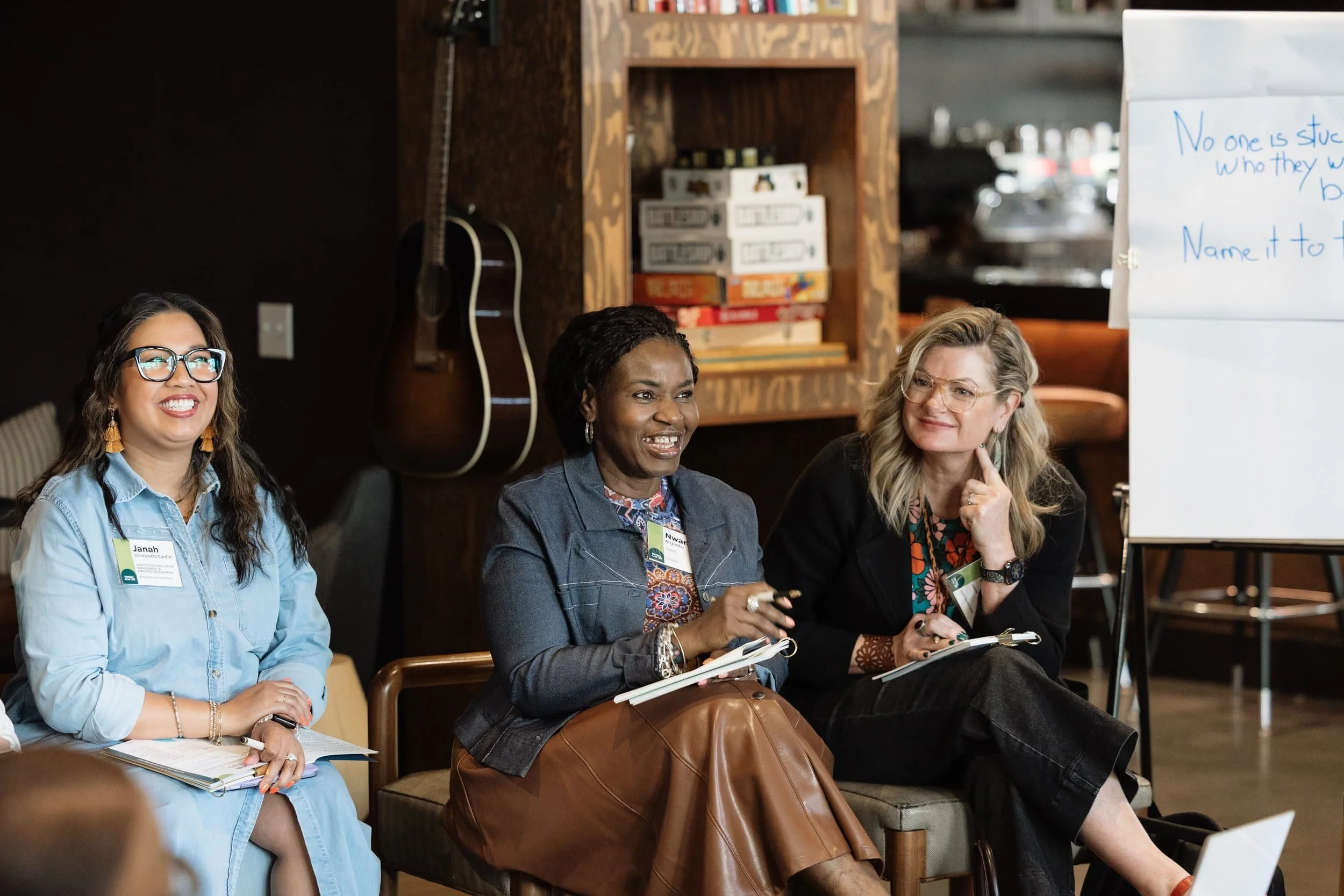 Three women smile during a casual meeting at a leadership conference in Seattle, with notebooks, a whiteboard, and guitar behind them.