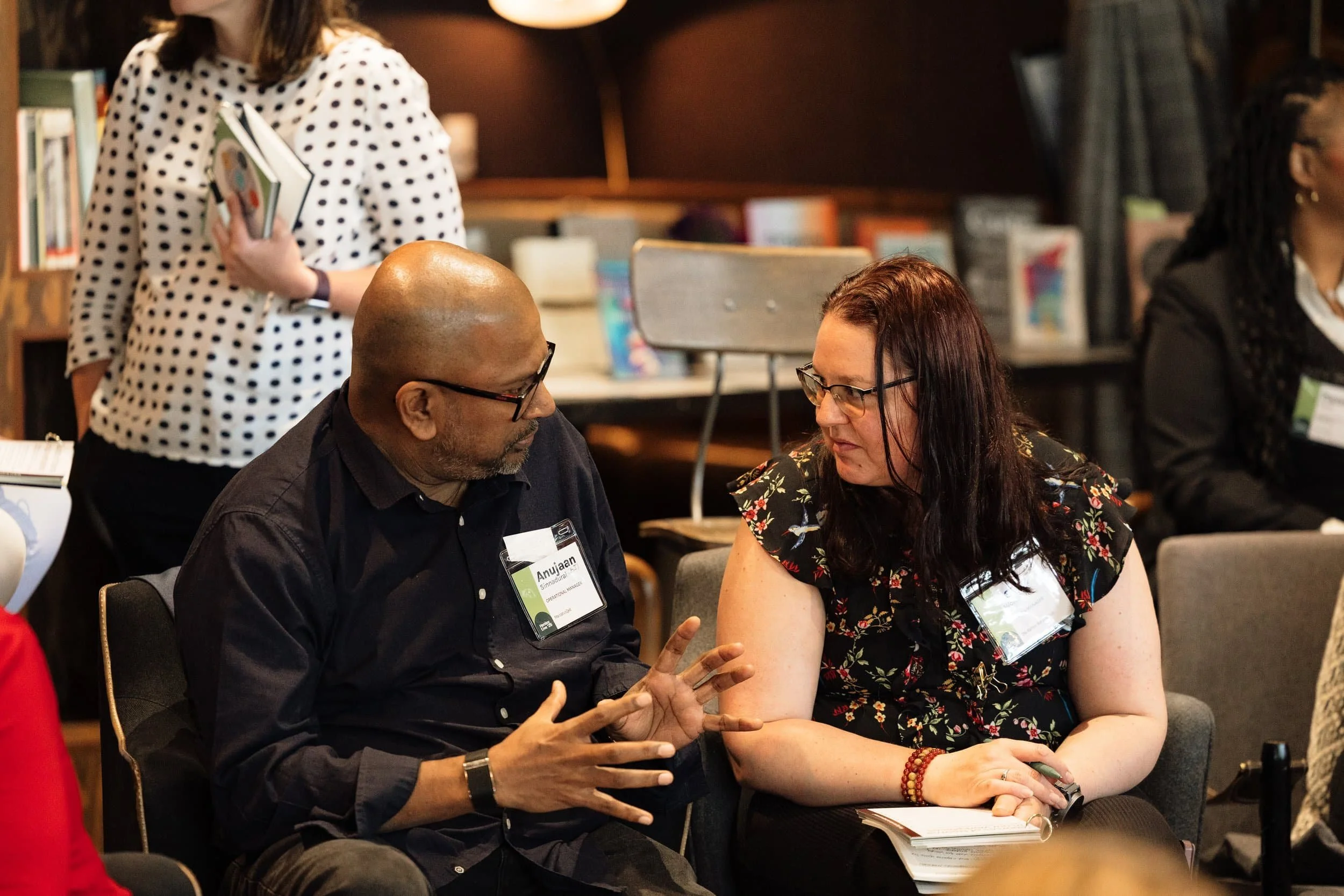 Two people chat at the Seattle Leadership Conference, both wearing name tags, with bookshelves and attendees in the background.