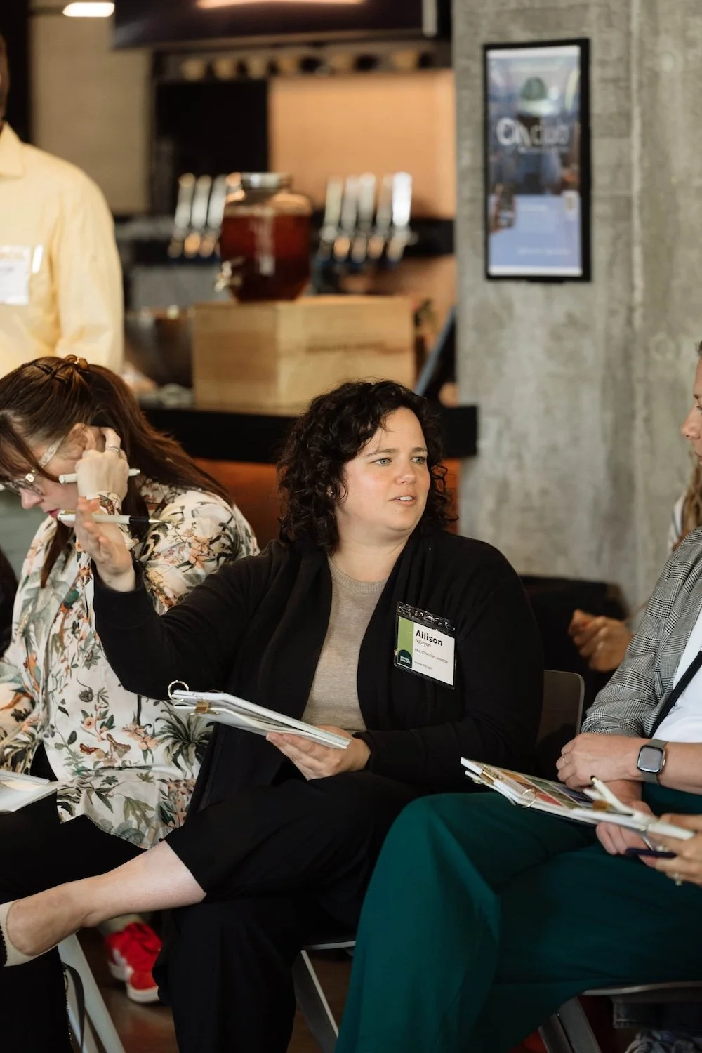 At a PNW business conference, a woman with curly hair speaks to the group, holding papers and gesturing as she shares ideas.
