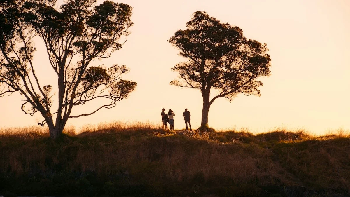 A small group stands together on a grassy hillside at sunset, suggesting connection, shared purpose, and community.