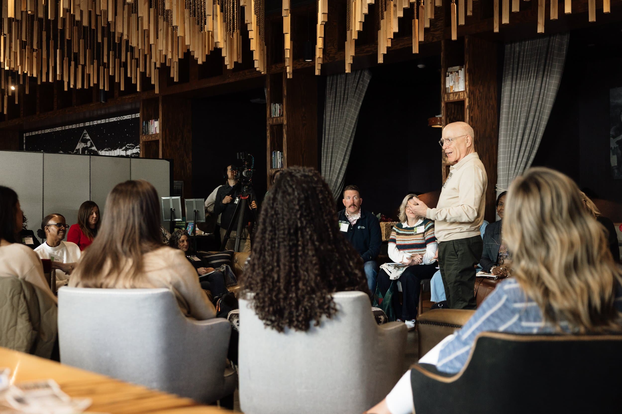 A man leads a business conference on PNW trends, speaking to a seated group in a modern room with wooden decor and hanging lights.