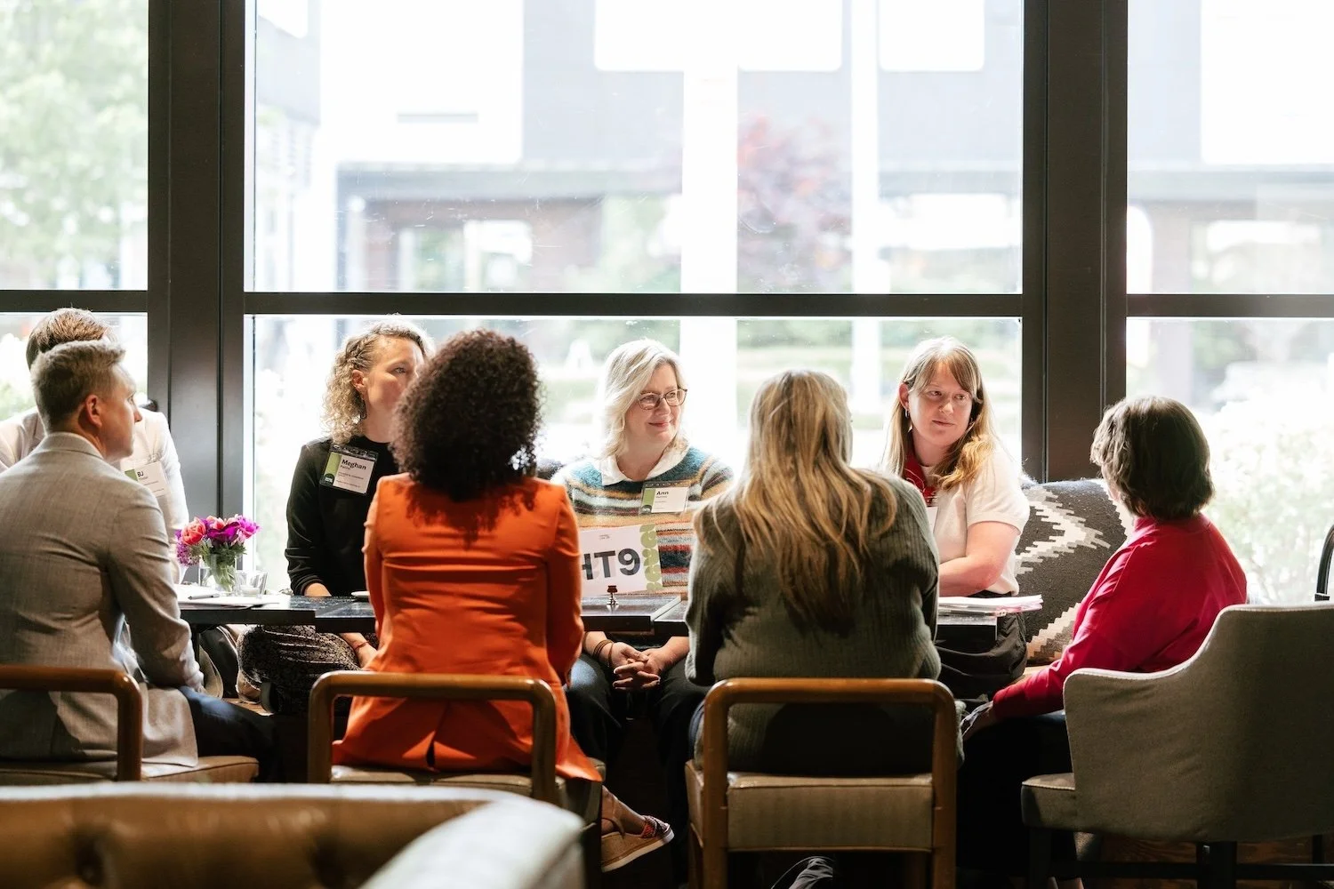 A group of people sit in a circle talking near large windows in a bright, modern room at Henley Live.