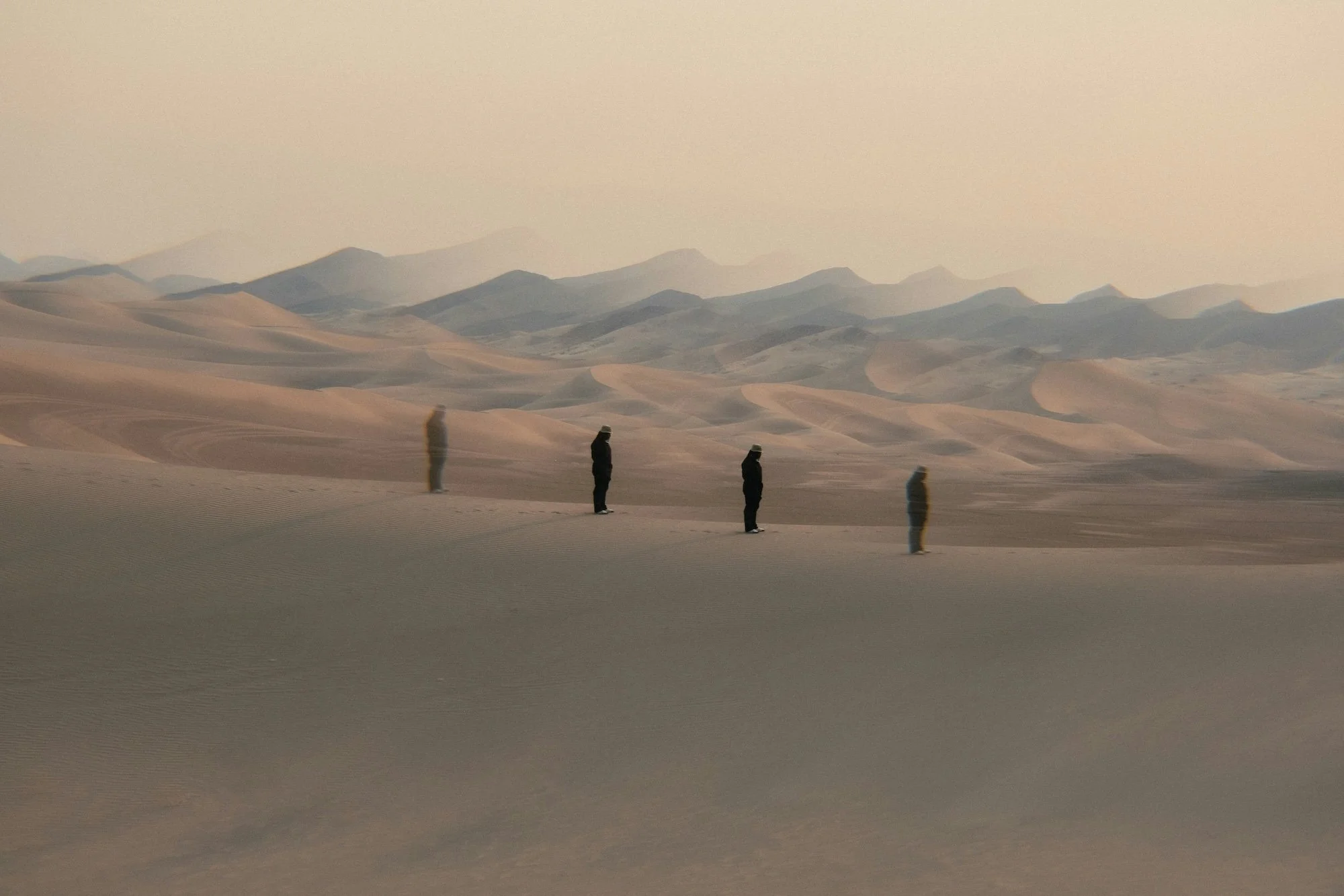 A lone figure repeated across a sand dune in a wide desert scene with rolling dunes and distant mountains.