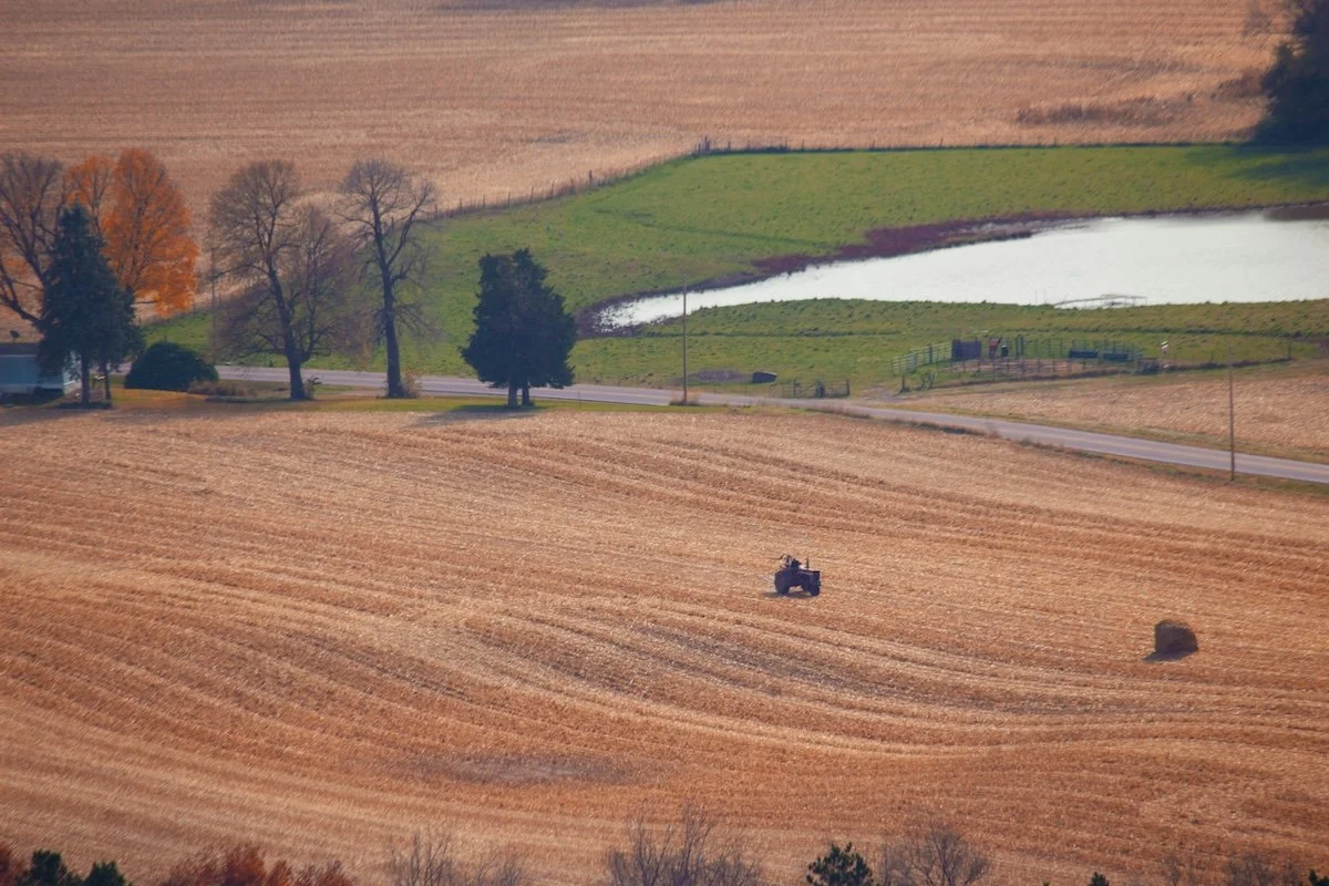 Autumn farmland with gently curving rows of harvested fields, a small tractor, rolling green hills, and a pond in the distance.