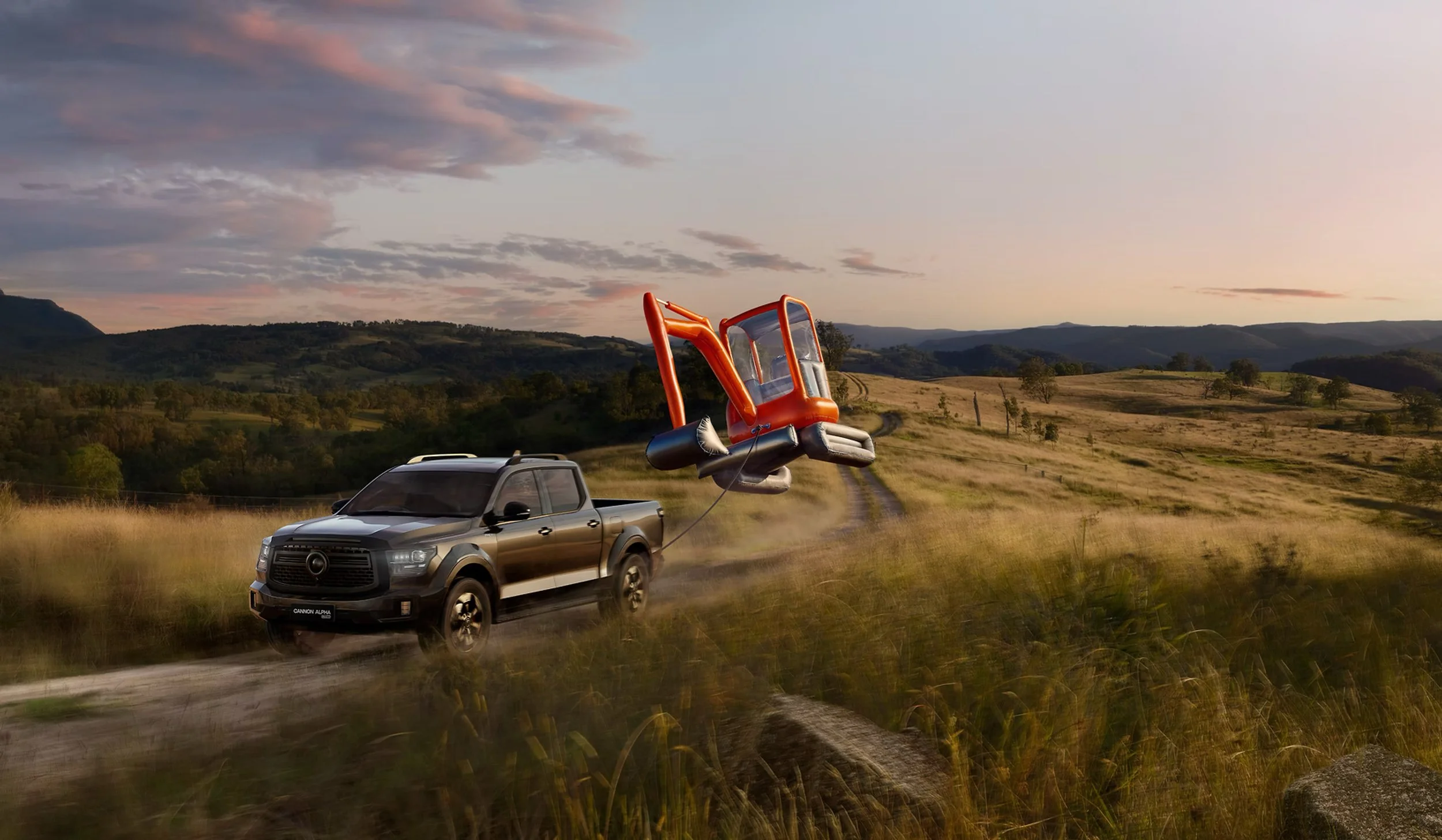 A black pickup truck towing an inflatable orange and grey helicopter on a dirt road in a grassy field at sunset.