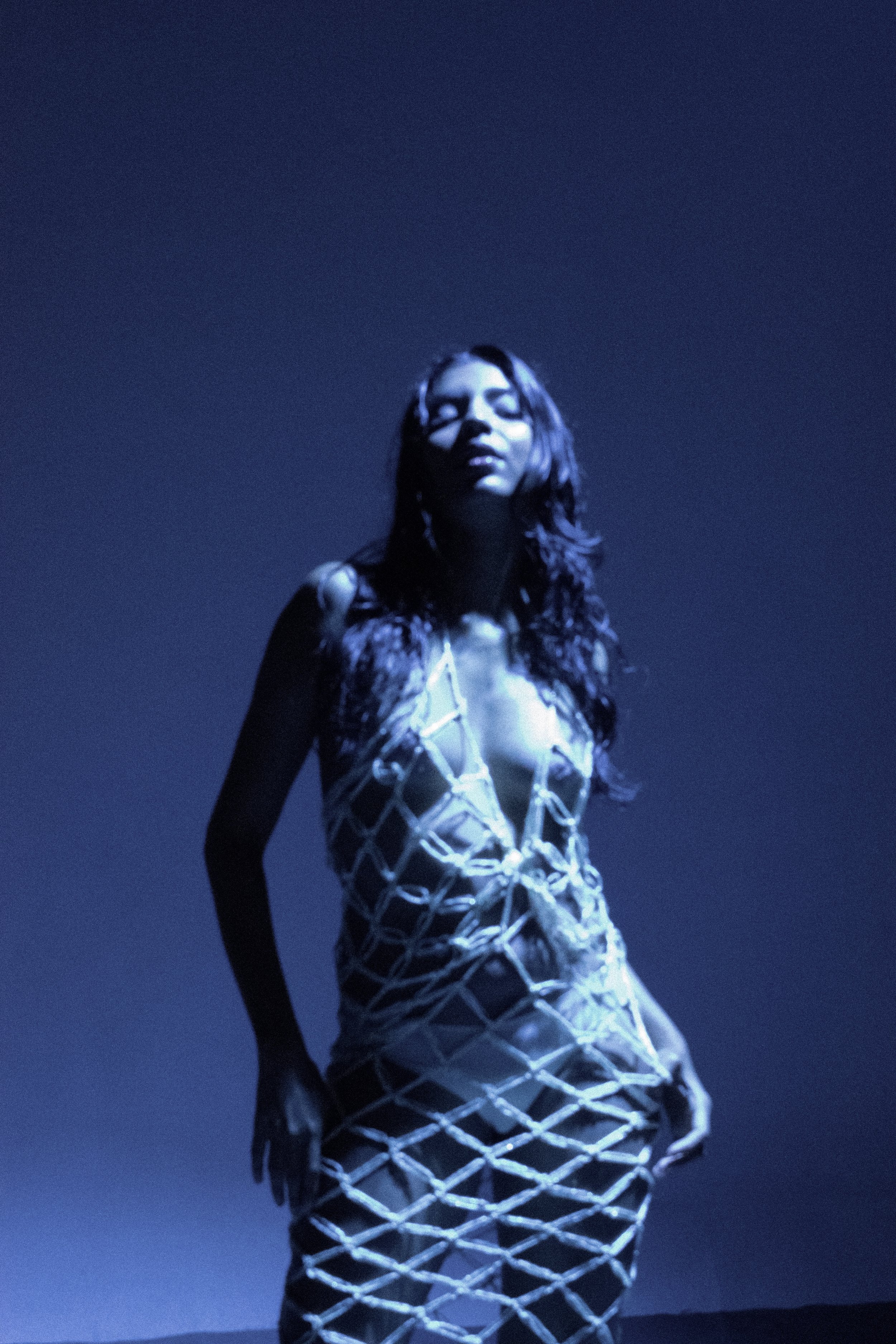 A woman with long, wavy hair wearing a revealing dress made of netting or ropes stands against a dark blue background, illuminated with dramatic lighting.