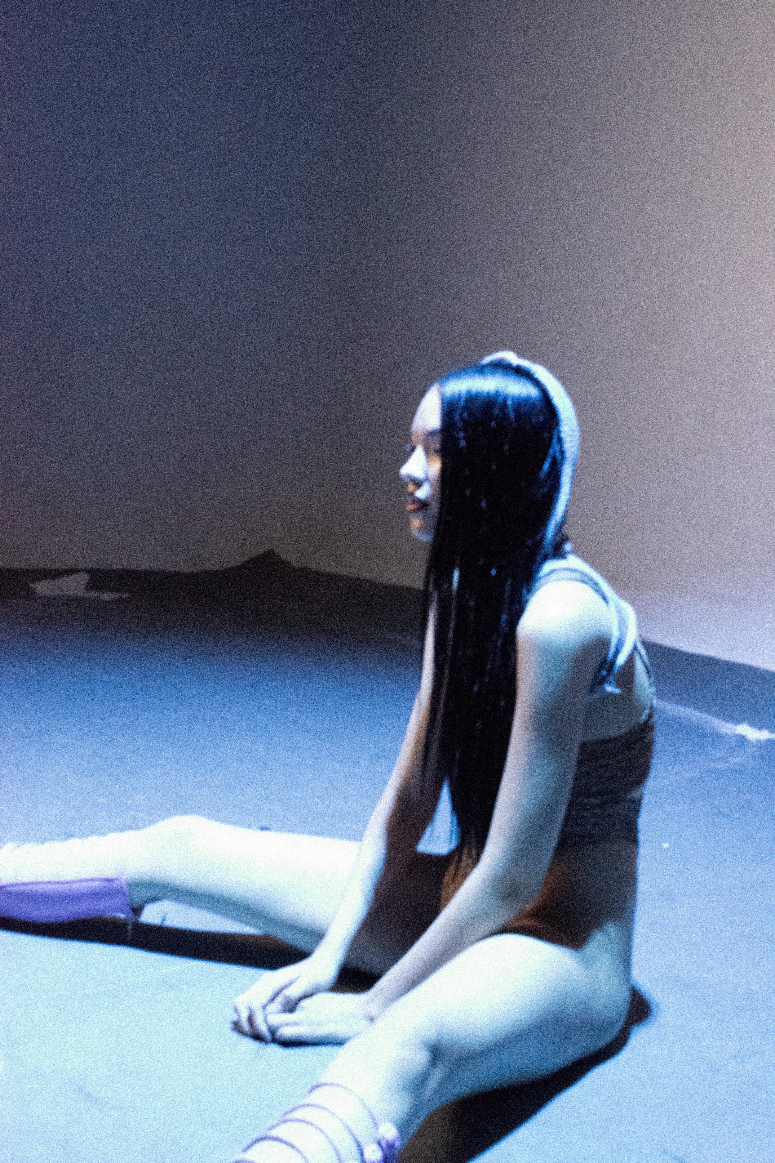 A woman with long black hair is sitting on the ground with her legs stretched out, wearing a dark tank top and colorful striped socks, against a dark background with blue lighting.
