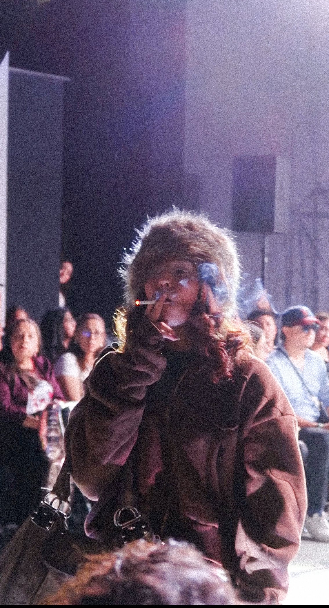 A young woman with curly hair wearing a brown jacket, smoking a cigarette, and holding a spray can in her hand at a crowded event or concert.