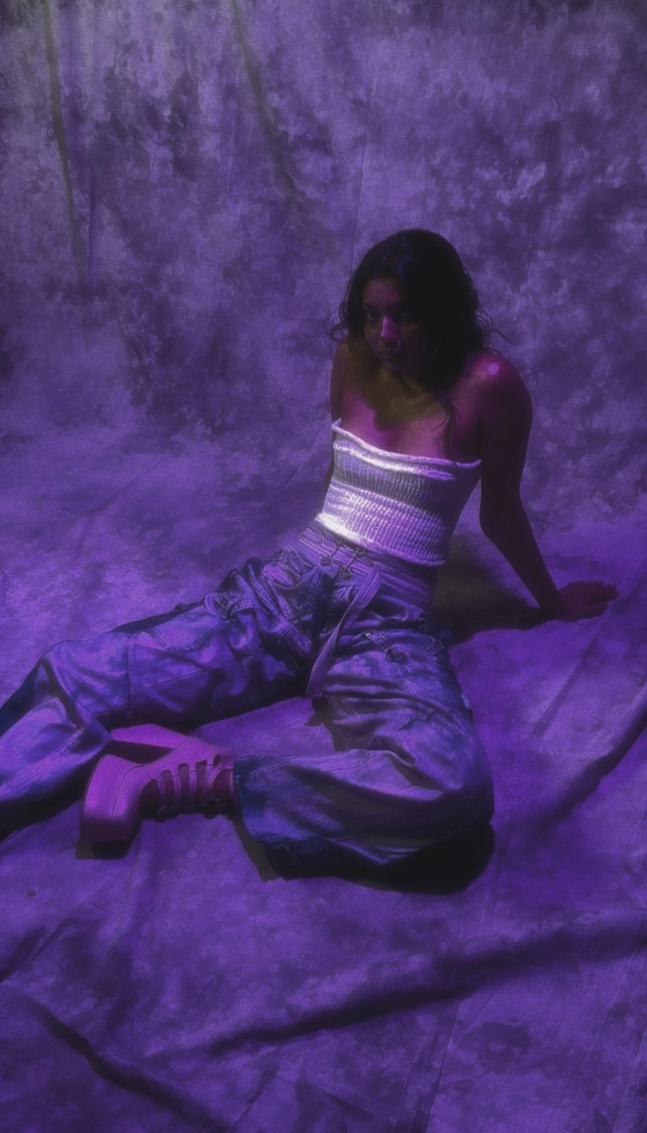 A woman with dark, wavy hair sitting on the floor against a textured purple wall. She wears a white cropped top, baggy jeans, and pink shoes, with purple lighting creating a moody atmosphere.