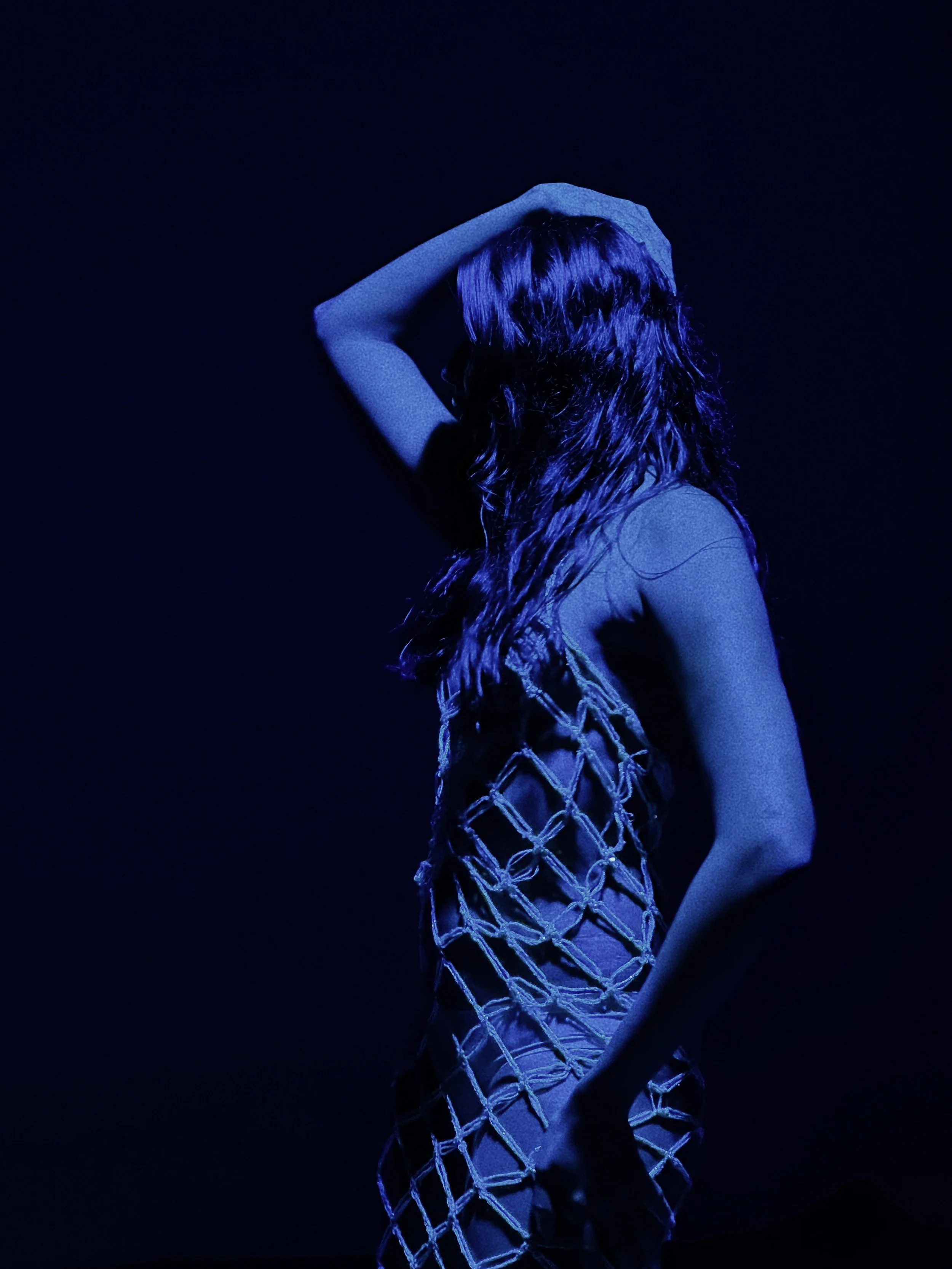 A woman with long, wavy hair is posing in a dark setting, illuminated by blue lighting, wearing a dress made of net-like material.
