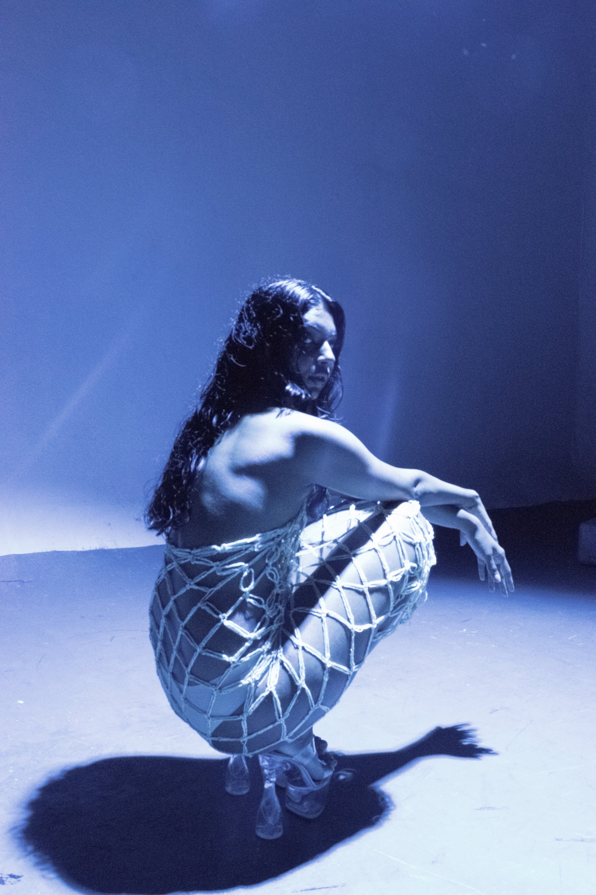 A woman with wet hair crouches on the floor, wearing a white netted dress, under blue stage lighting.
