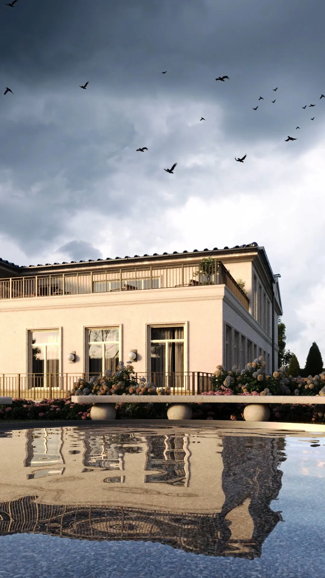 A white building with large windows and a balcony, surrounded by flowers, with a reflective surface in the foreground showing the building and a flock of birds flying in a cloudy sky.