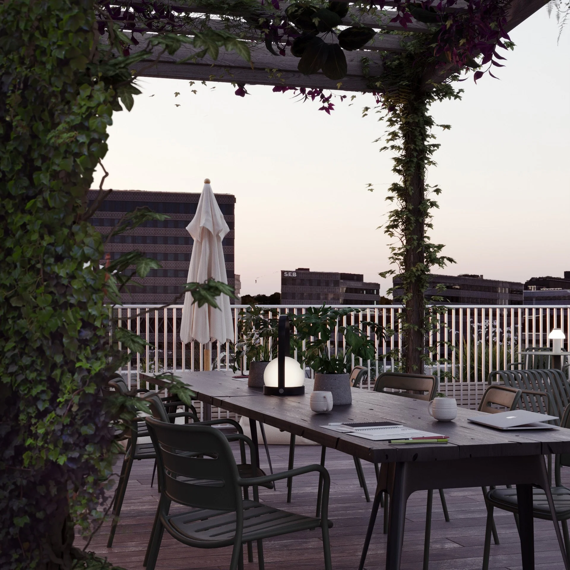 An outdoor dining area with a long table, chairs, and a white umbrella, surrounded by greenery and city buildings in the background during dusk.