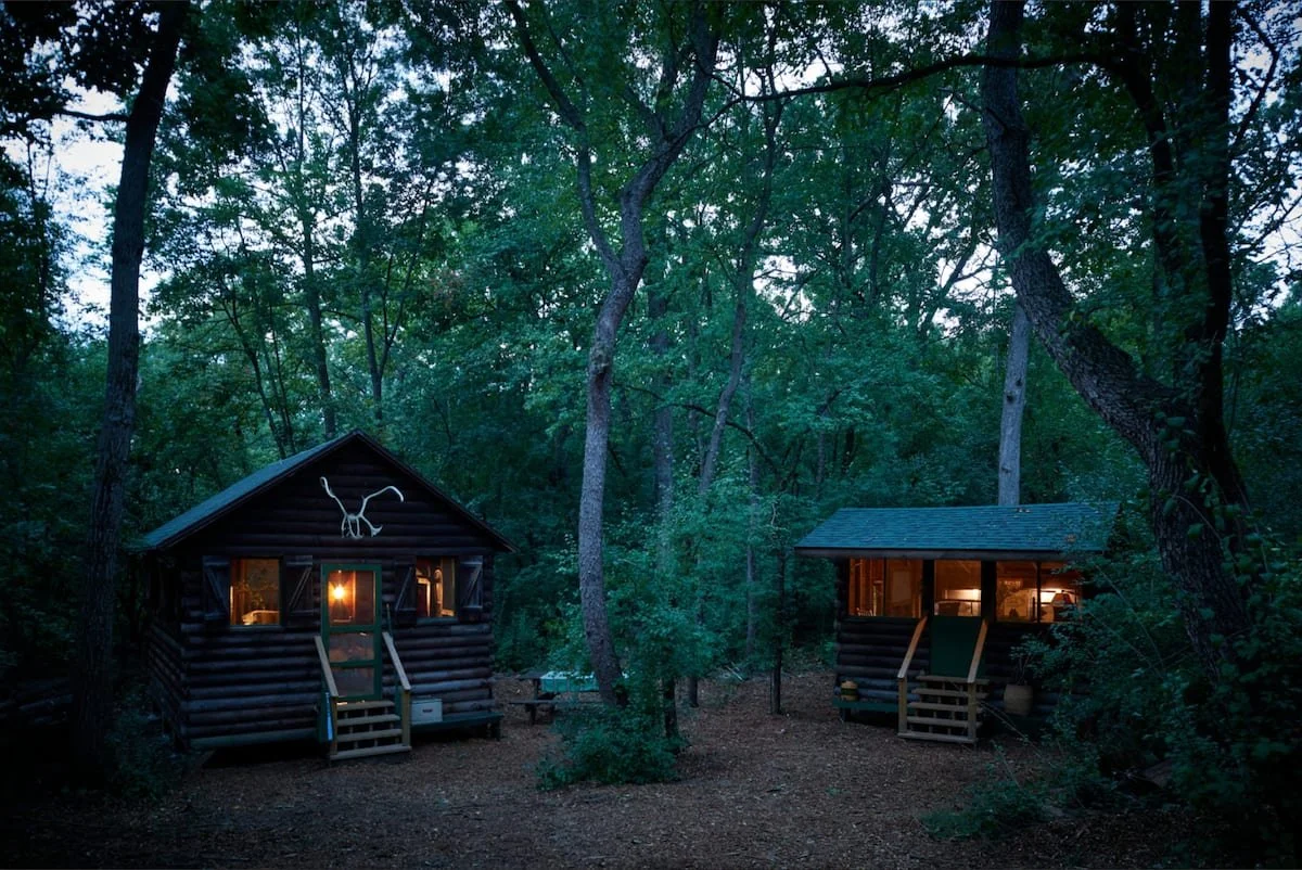 Image of Writers in the Dark. Sun is setting in the sky, there are a lot of green trees, and two cabins with their lights on..