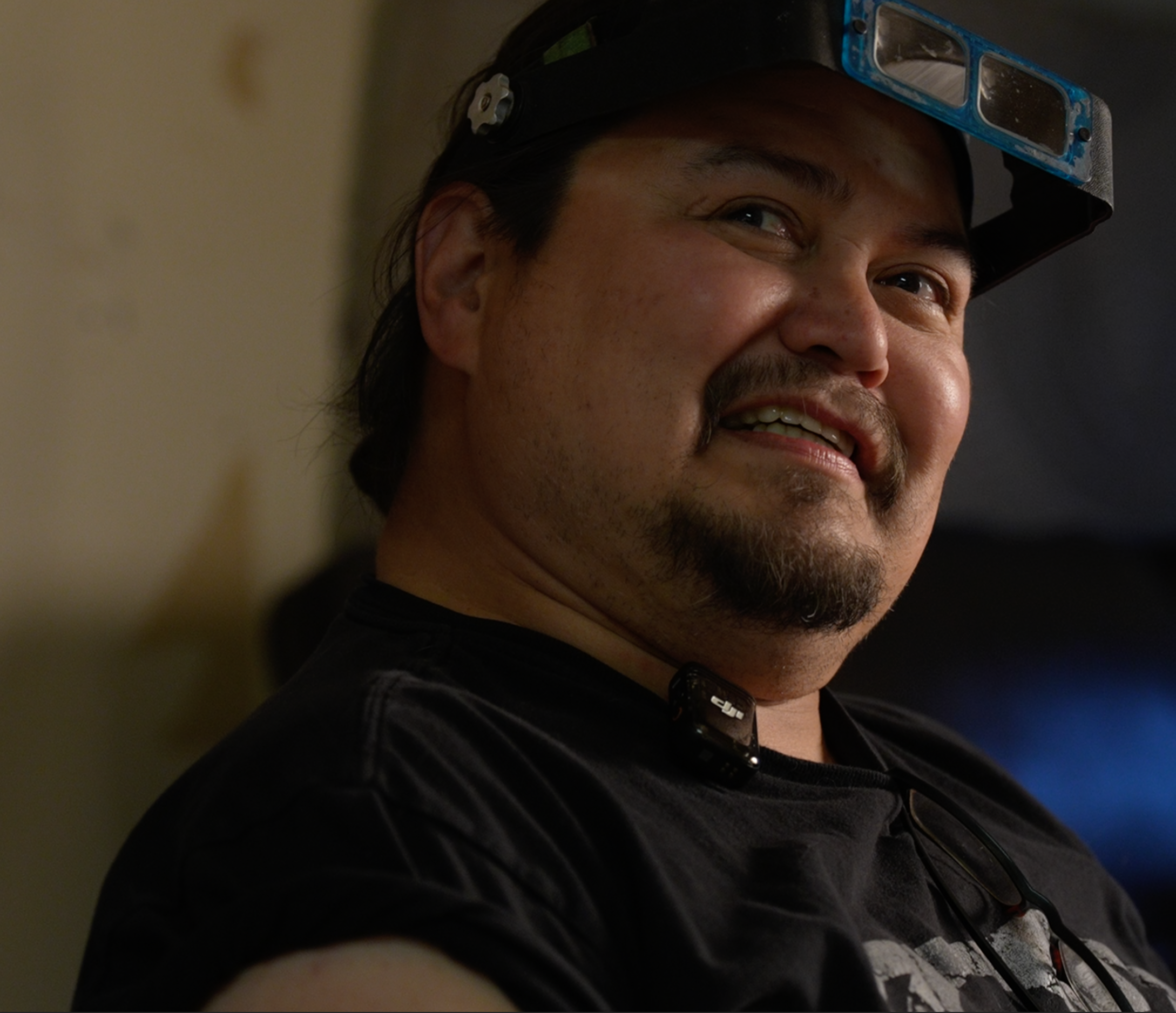 A close up headshot of Thomas Fawcett, a Carcross Tagish First Nation (CTFN) Carver and Metalworker. He is wearing a specialized head mounted magnifying glasses used for intricate carving and a black tshirt with a small lapel microphone.