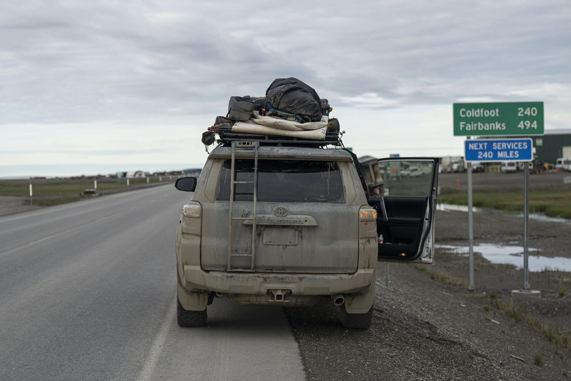 Rear view of a rugged SUV with camping gear strapped to the roof, on a two-lane highway near Coldfoot, Alaska. A green road sign shows Coldfoot 240 miles ahead, under a cloudy sky.